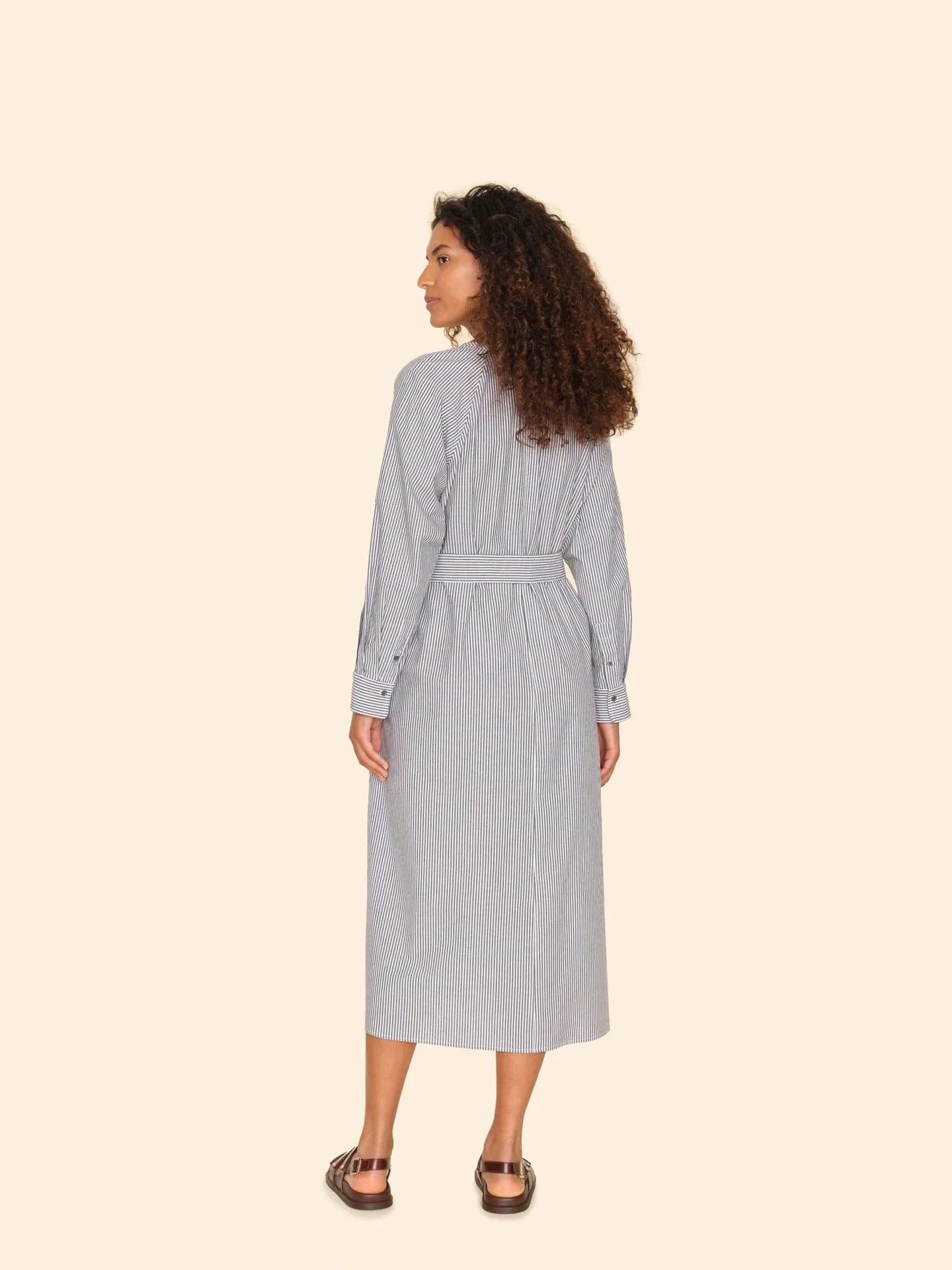 A person with curly hair stands with their back to the camera, wearing the Sylvie Dress by Xirena, characterized by its blouson sleeves and striped midi design. Paired with sandals, the cotton pencil stripe fabric of the dress gracefully flows down to ankle-length against a solid light-colored background.