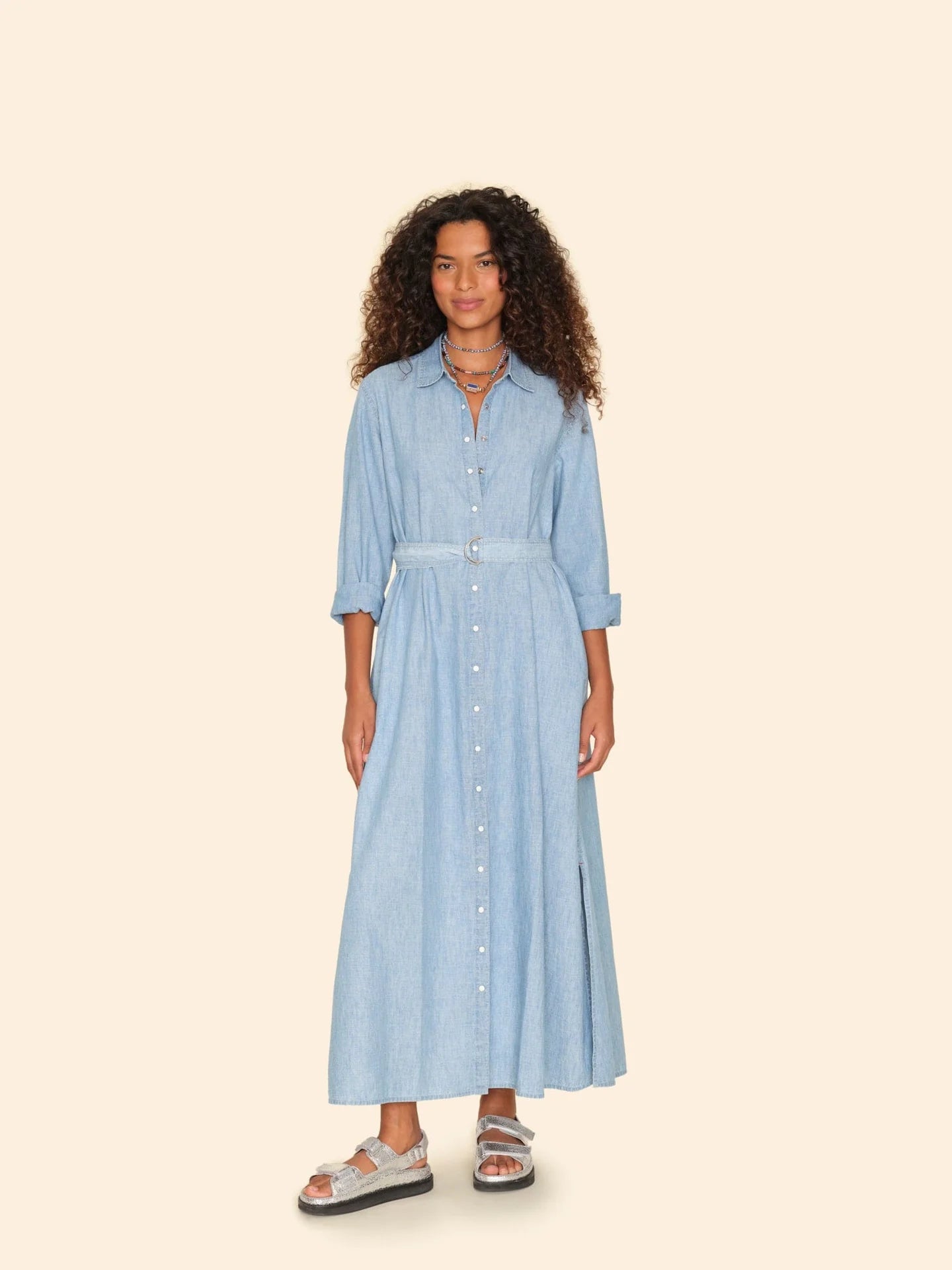 A woman wearing the chic Bowen Dress by Xirena, featuring pearlized snap buttons and a belt, stands against a beige background. Her maxi A-line ensemble, paired with silver sandals, beautifully complements her curly hair and layered necklaces.