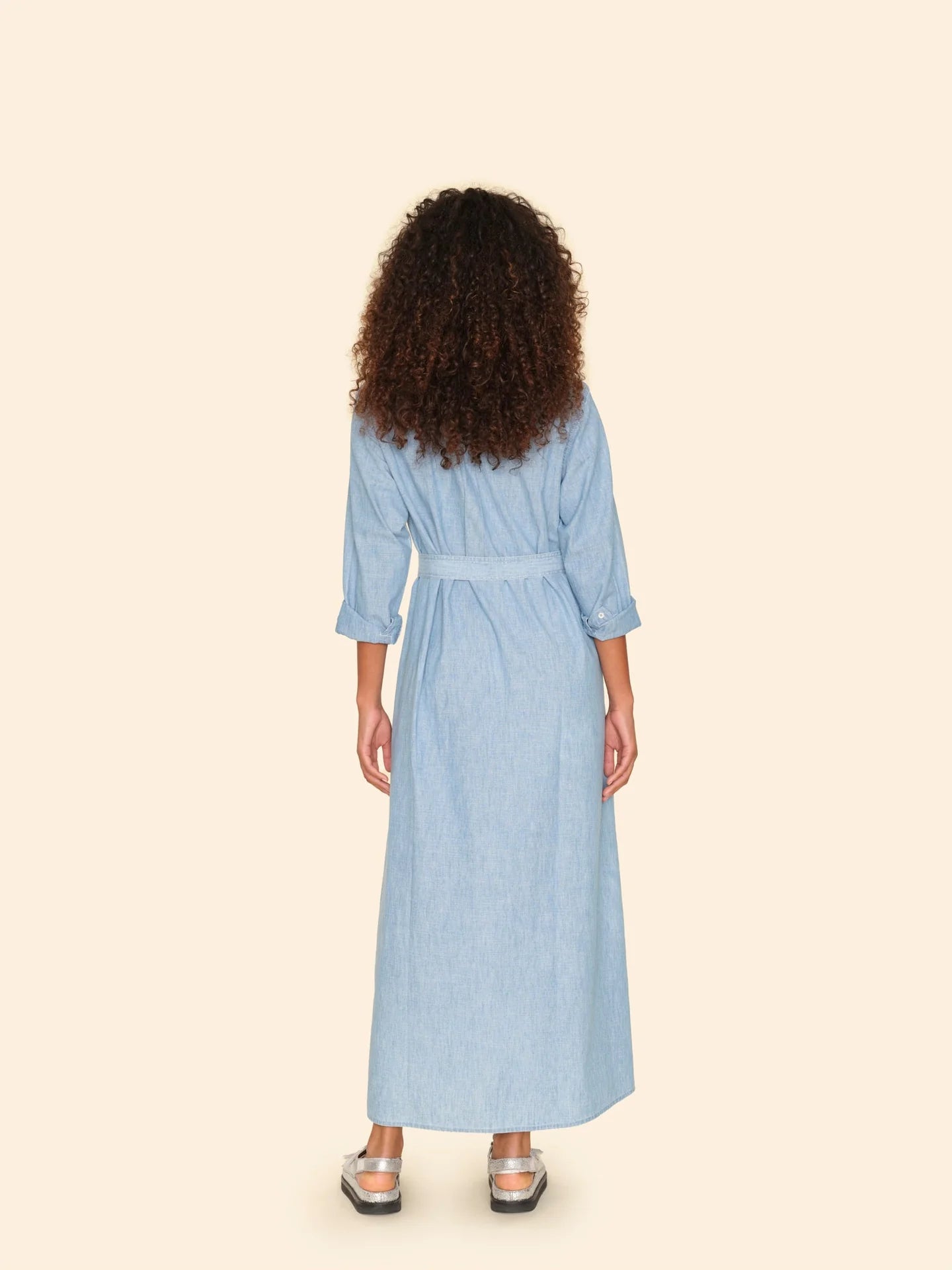 A person with curly hair stands facing away, showcasing the Bowen Dress by Xirena, a light blue chambray design featuring pearlized snap buttons and a belt. The dress's three-quarter sleeves add elegance, perfectly complemented by silver sandals against a plain beige background.