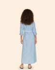 A person with curly hair stands facing away, showcasing the Bowen Dress by Xirena, a light blue chambray design featuring pearlized snap buttons and a belt. The dress's three-quarter sleeves add elegance, perfectly complemented by silver sandals against a plain beige background.