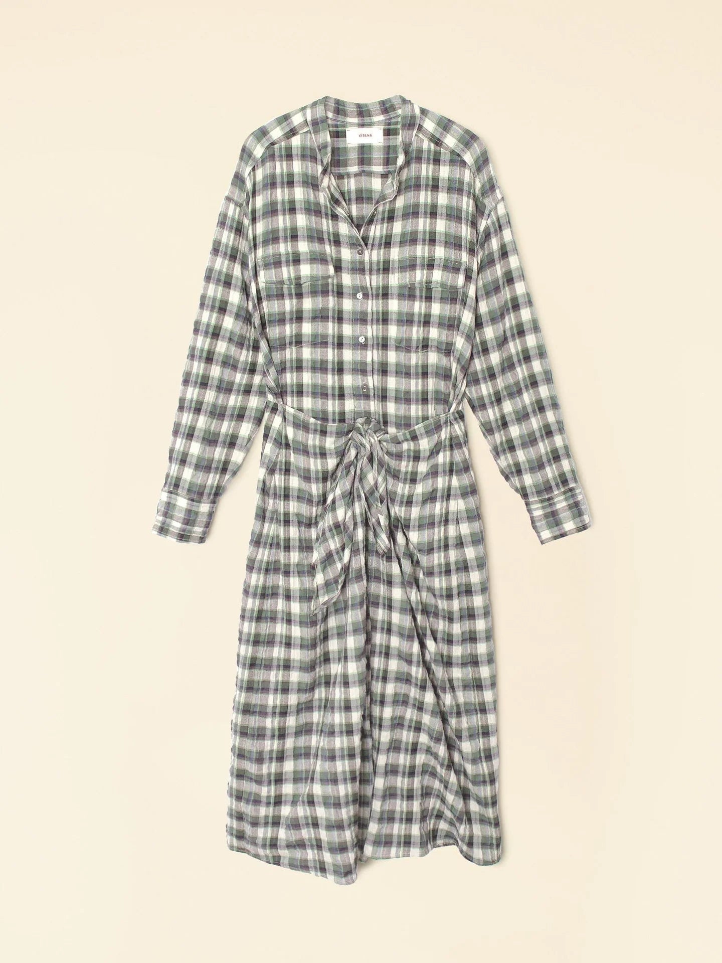 The Alanis Dress by Xirena is a midi shirtdress with a green, white, and black checkered pattern. It features long sleeves, a button-front design, and a waist tie, all displayed on a light background.