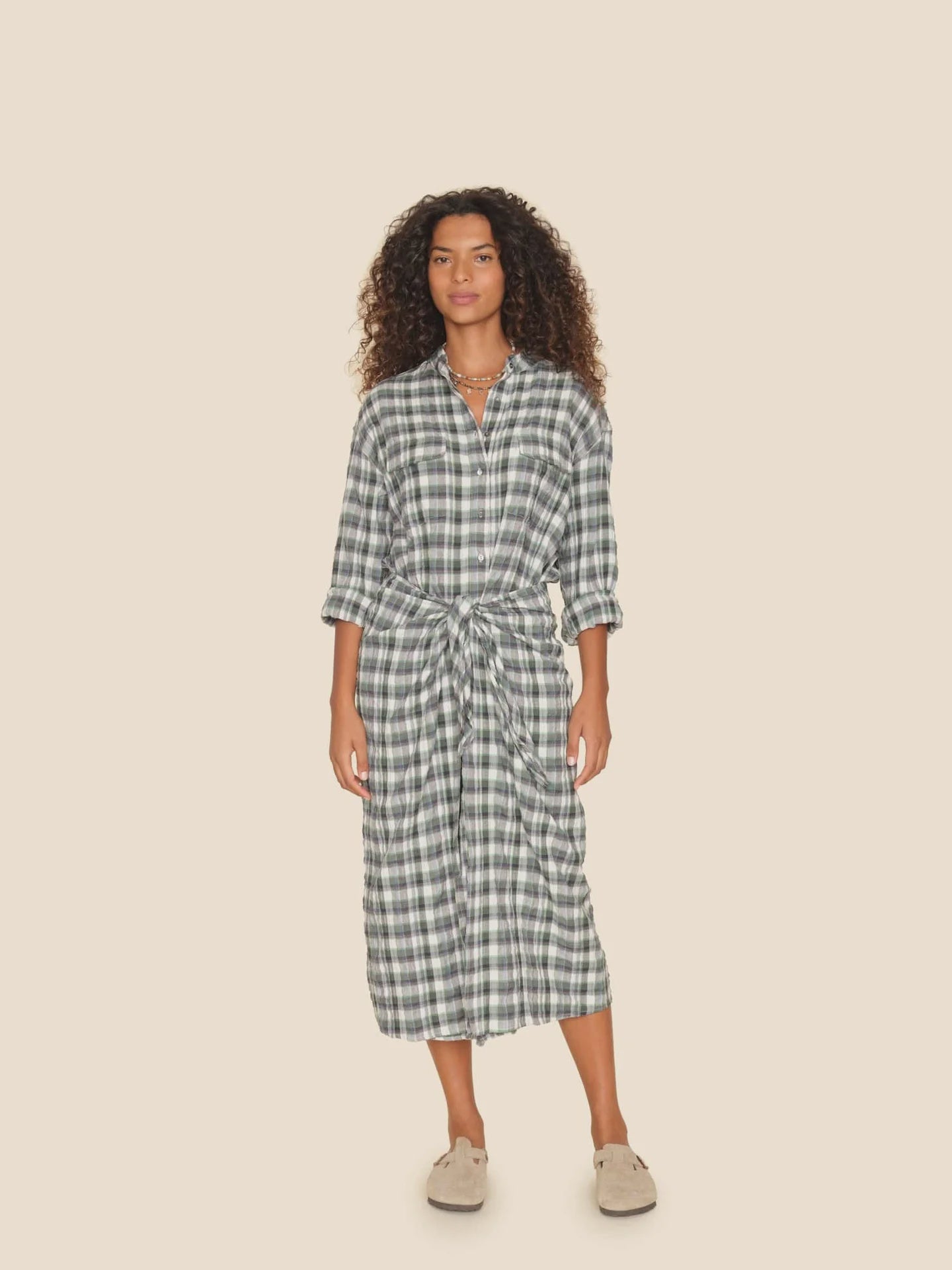 Against a beige backdrop, a person with curly hair sports the Alanis Dress from Xirena, showcasing long sleeves and a self-tie wrap waist in a green and white plaid pattern, perfectly paired with beige shoes.