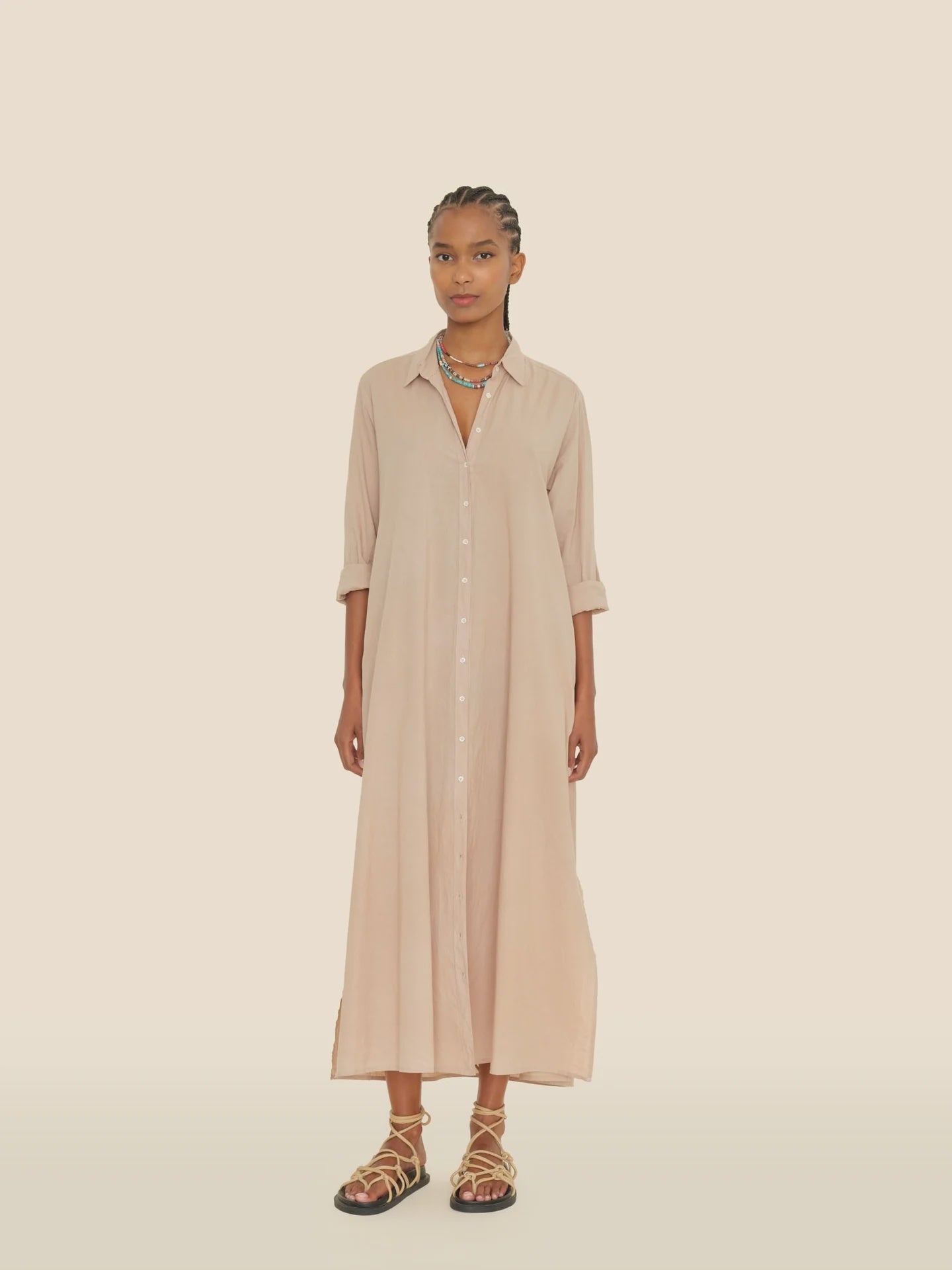 In front of a beige backdrop, an individual models a Xirena dress fashioned from soft cotton poplin. The Boden dress features rolled-up sleeves, and they accentuate their look with braided hair, layered necklaces, and intricately laced-up sandals.