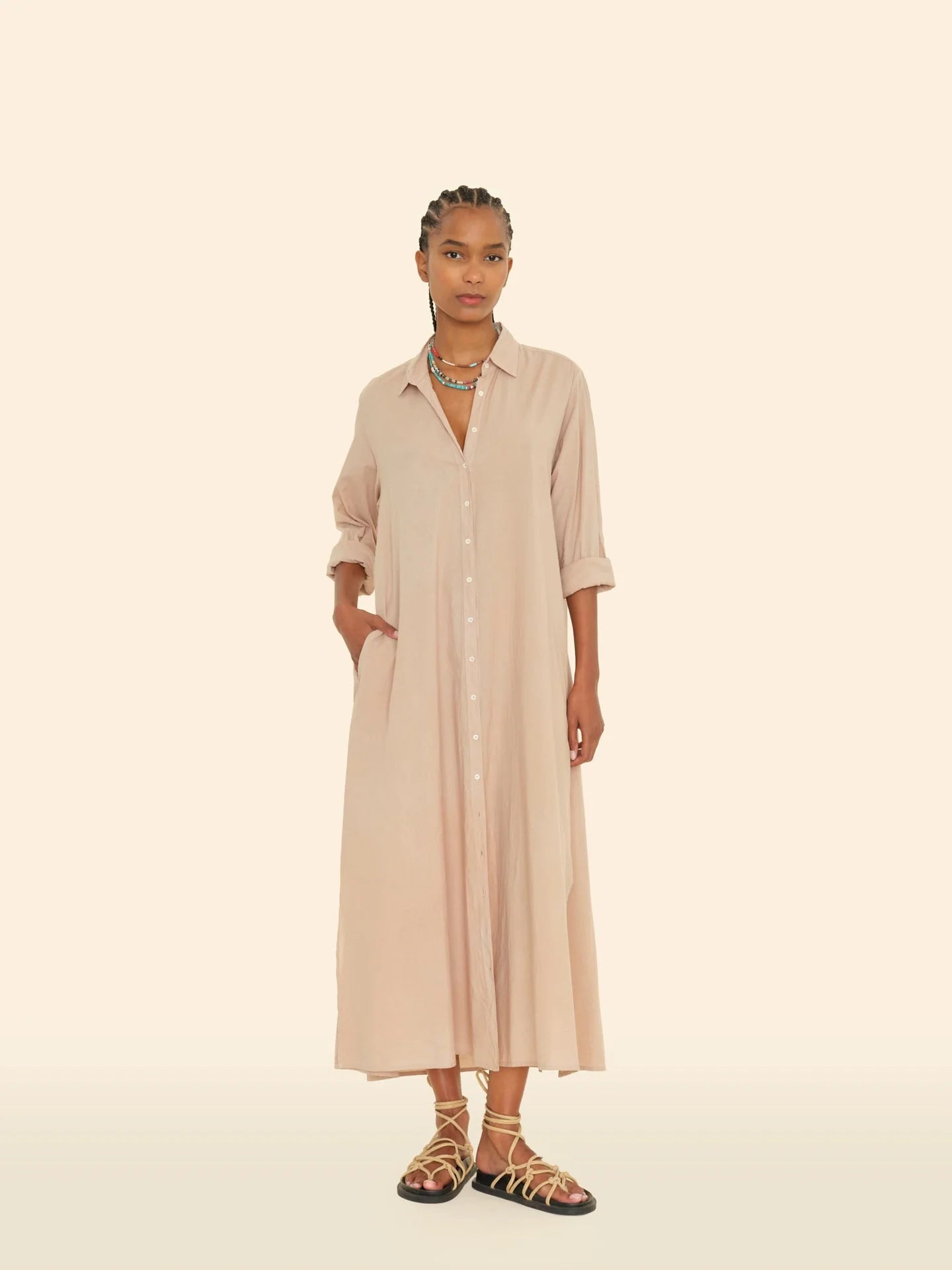 A person stands against a light beige background, wearing the Boden Dress by Xirena, a long button-front tailored shirtdress in light tan with sleeves rolled to the elbows. They have braided hair, layered necklaces, and strappy sandals.