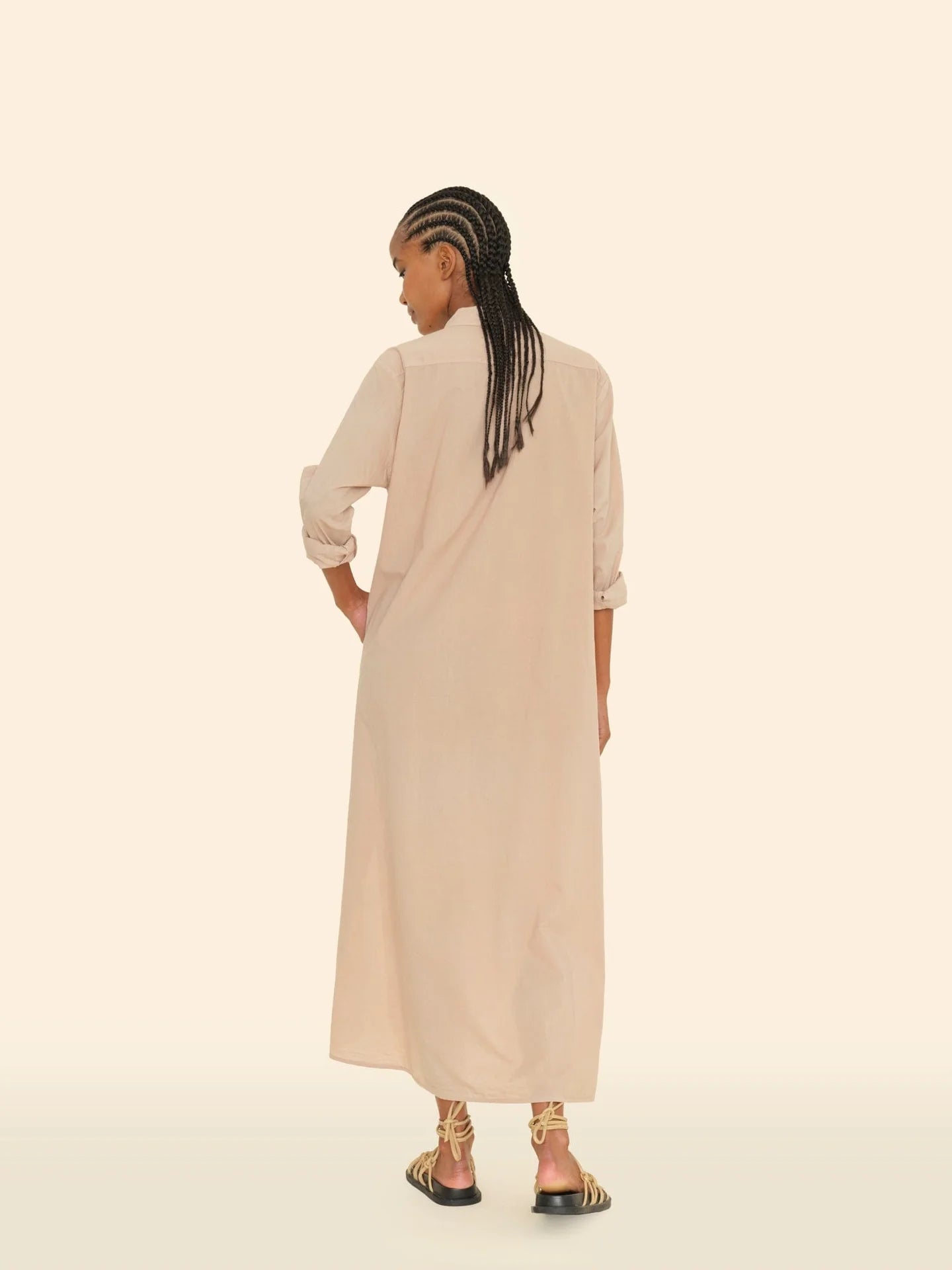 A person with braided hair stands facing away, wearing the Boden Dress, a long, tailored piece by Xirena, paired with strappy sandals. The background is a simple off-white color.