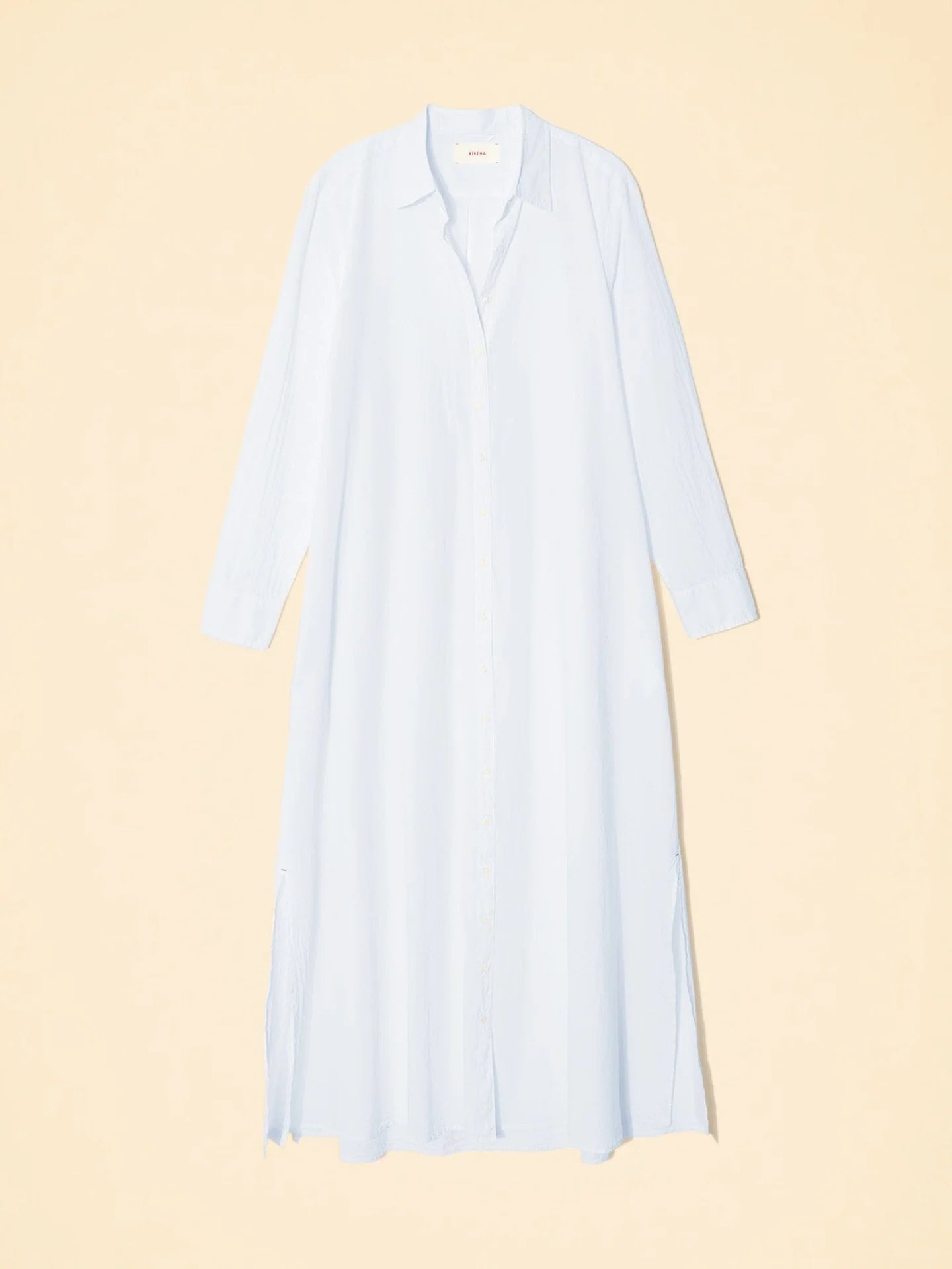 The Boden Dress by Xirena showcases a long light blue classic shirtdress with a collar and long sleeves, set against a beige background. Button fastenings enhance the tailored fit, while side slits at the hem add elegance to this cotton poplin design.