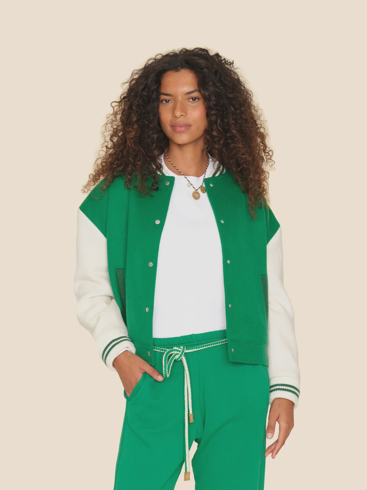 A person with curly hair confidently poses in a Xirena Varsity Jacket, featuring green with white sleeves, paired with matching green pants against a plain beige background.