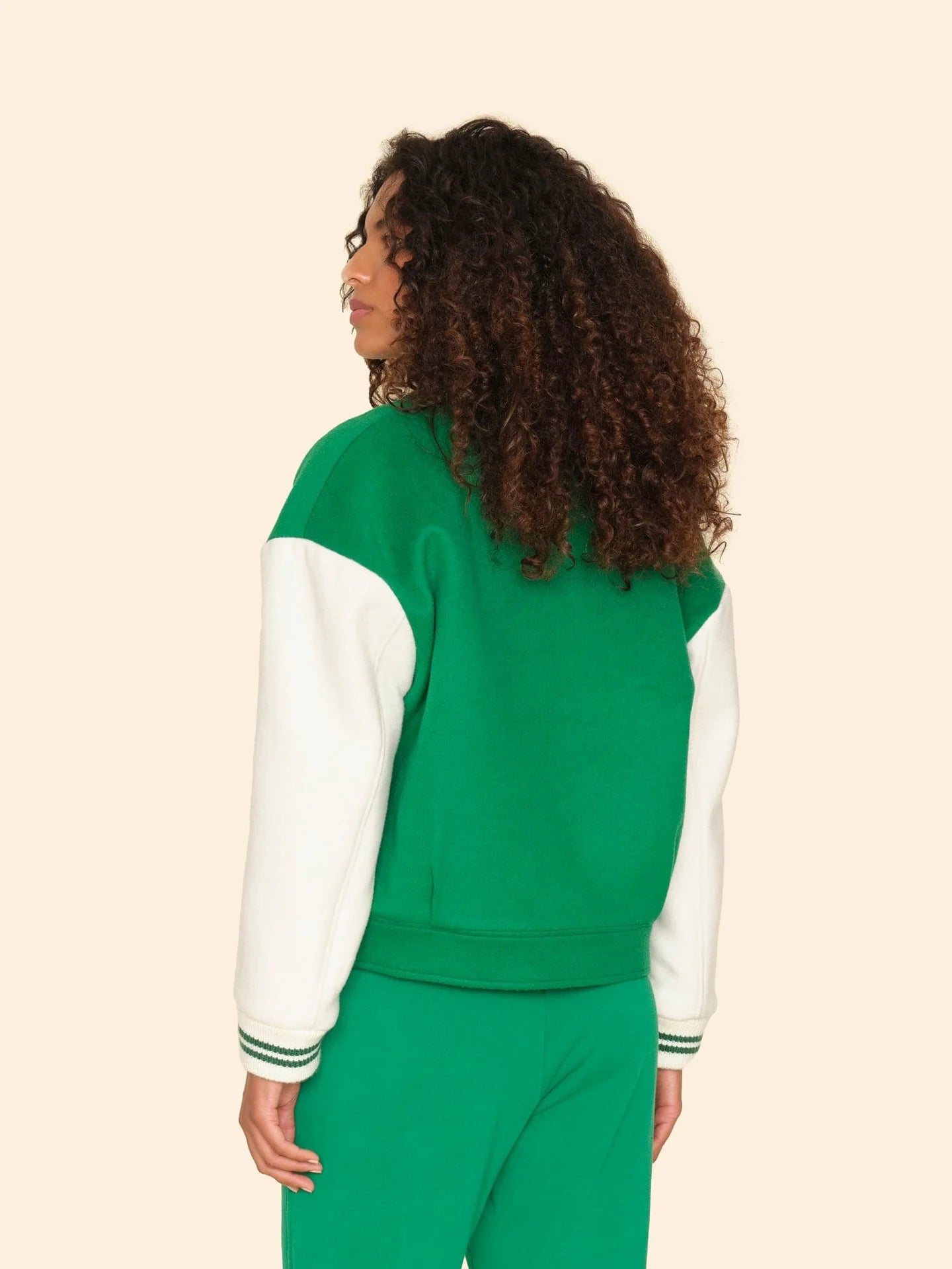 A curly-haired person, wearing a green and white wool blend varsity jacket by Xirena with matching green pants, stands with their back to the camera against a beige backdrop.