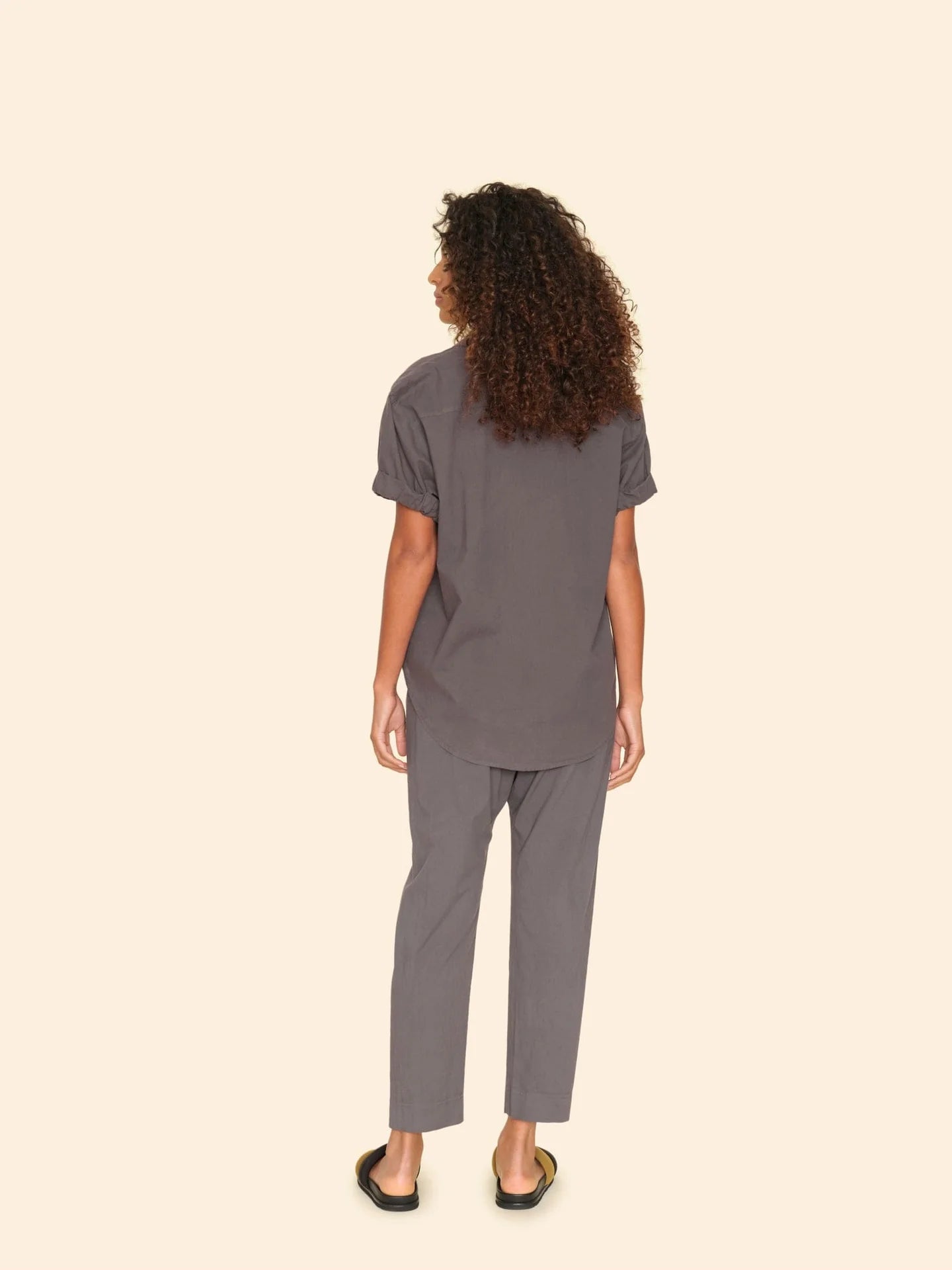 A person with curly hair stands facing away from the camera, styled in a gray cotton poplin short-sleeve shirt and matching Xirena Draper Pant Ash with a drawstring waist. Black sandals complete their look, and they are set against a plain beige background.