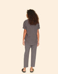 A person with curly hair stands facing away from the camera, styled in a gray cotton poplin short-sleeve shirt and matching Xirena Draper Pant Ash with a drawstring waist. Black sandals complete their look, and they are set against a plain beige background.