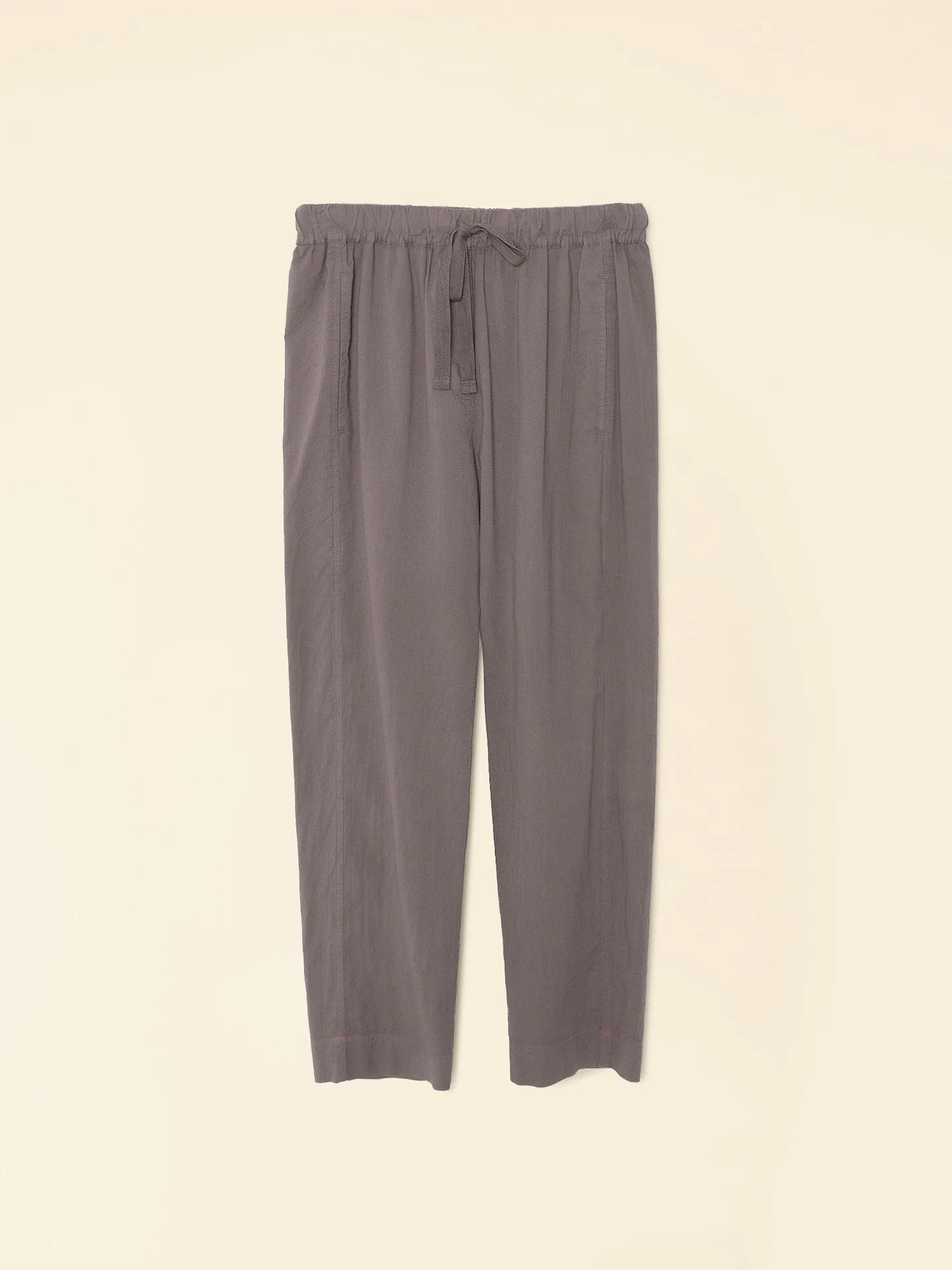 The Draper Pant Ash by Xirena is a pair of gray high-waisted pull-on pants with a drawstring waist, made from soft cotton poplin. They offer a wide, relaxed fit and are equipped with two side pockets, beautifully showcased on a plain beige background.