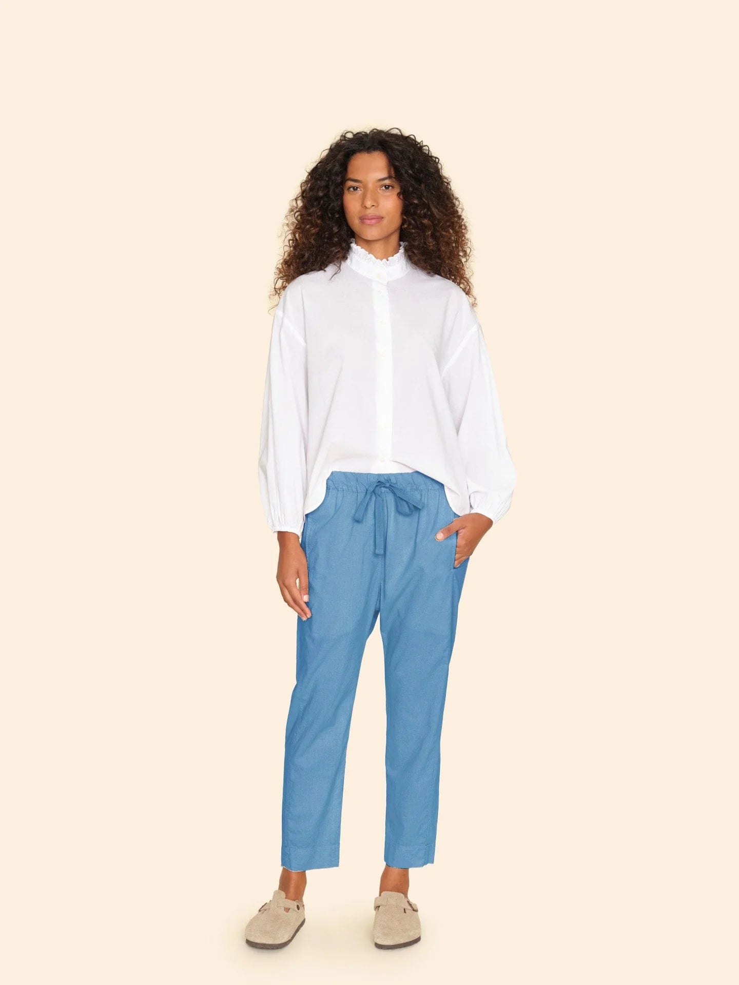 A person with long, curly hair wears a white blouse and Xirena's Draper Pant, featuring a drawstring waist. They stand against a plain, light background in this casual and relaxed ensemble.