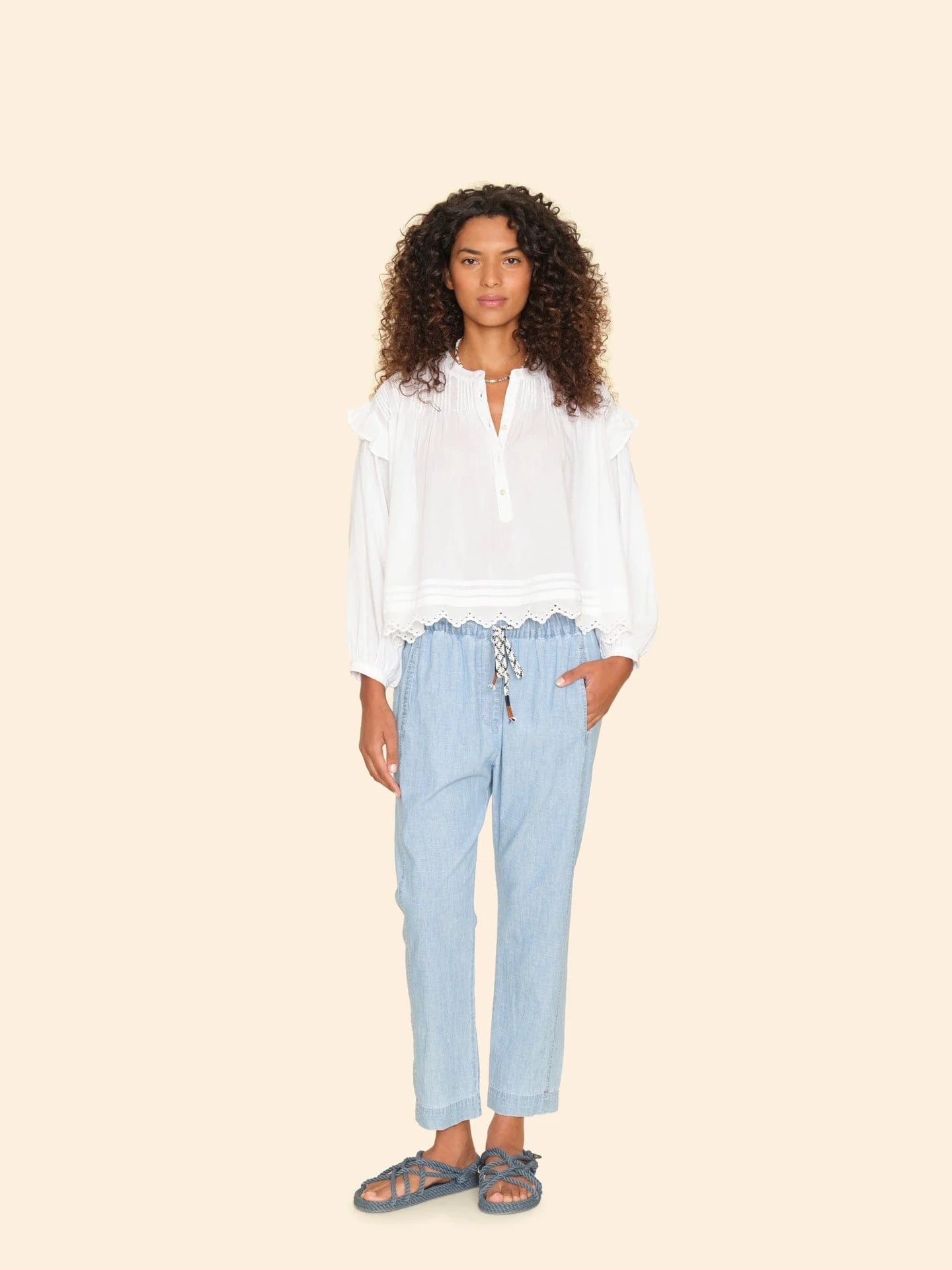 A person with curly hair stands against a plain background, wearing a white long-sleeve blouse and the Draper Pant by Xirena, featuring light blue pull-on style with a drawstring waist. Their right hand rests in a pocket, and they complete the look with dark sandals.