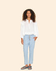 A person with curly hair stands against a plain background, wearing a white long-sleeve blouse and the Draper Pant by Xirena, featuring light blue pull-on style with a drawstring waist. Their right hand rests in a pocket, and they complete the look with dark sandals.