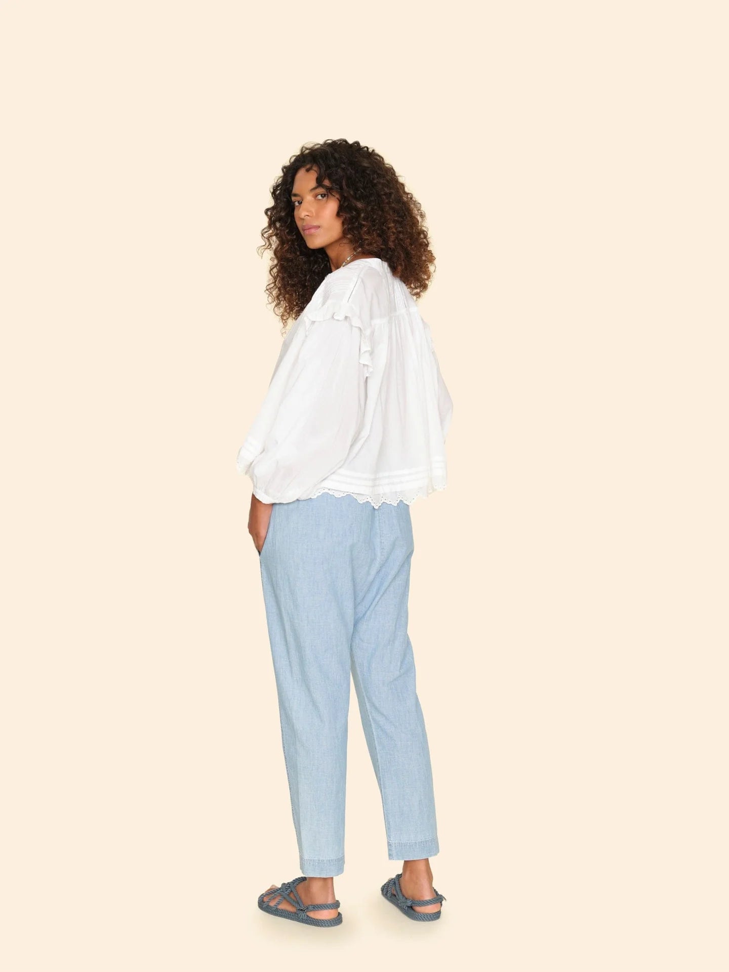 A woman with curly hair stands sideways in a white, ruffled blouse and wears Xirena's Draper Pant in light blue, featuring a drawstring waist. She casually rests her hands in her pockets and wears sandals against a plain beige background.