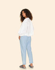 A woman with curly hair stands sideways in a white, ruffled blouse and wears Xirena's Draper Pant in light blue, featuring a drawstring waist. She casually rests her hands in her pockets and wears sandals against a plain beige background.