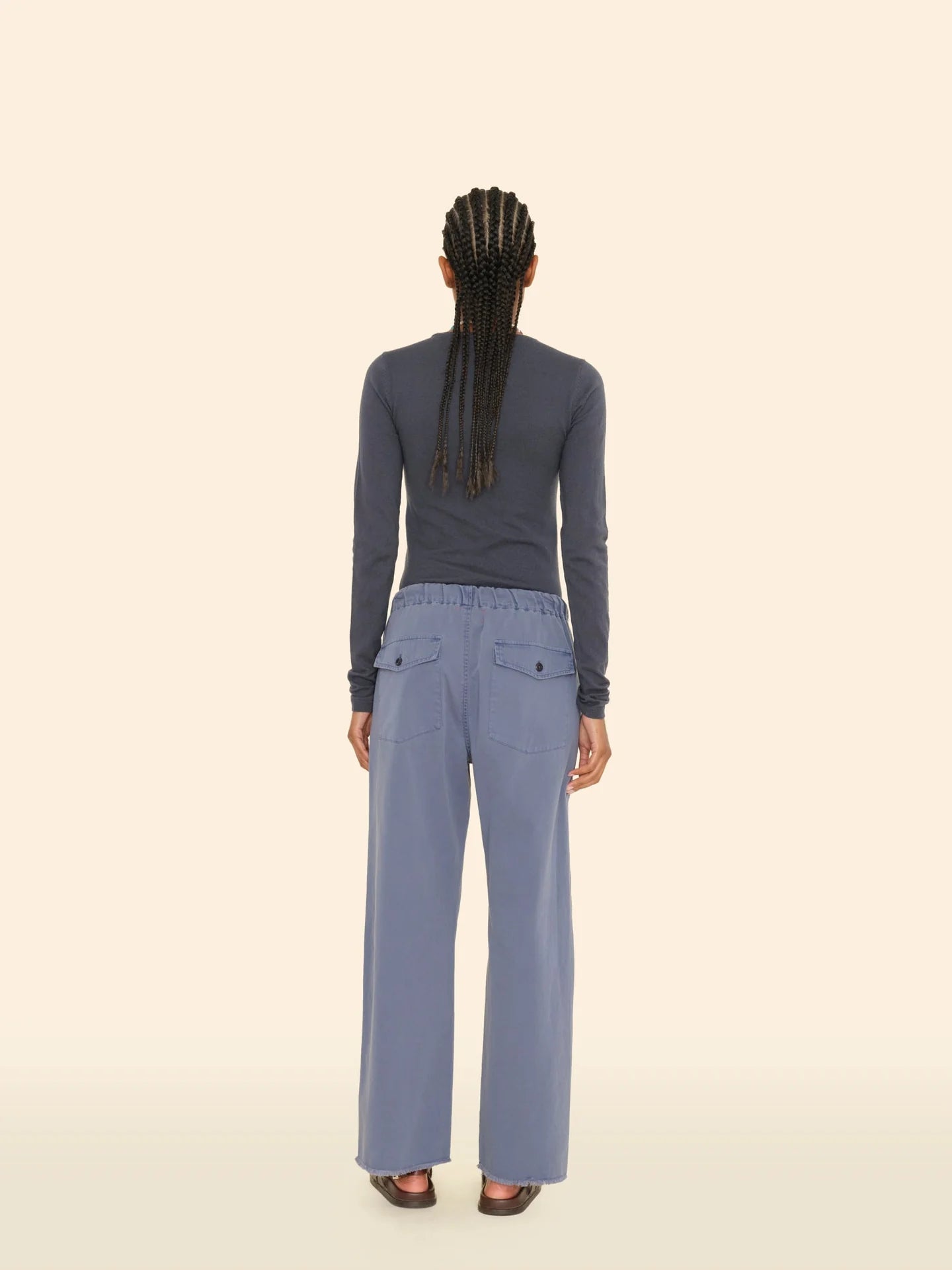 A person with long braided hair is shown from behind, wearing a dark long-sleeve top and the Eli Pant by Xirena in light blue cotton twill, featuring an elasticized waist. The backdrop is a plain, light beige color.