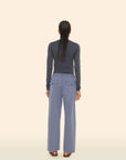 A person with long braided hair is shown from behind, wearing a dark long-sleeve top and the Eli Pant by Xirena in light blue cotton twill, featuring an elasticized waist. The backdrop is a plain, light beige color.