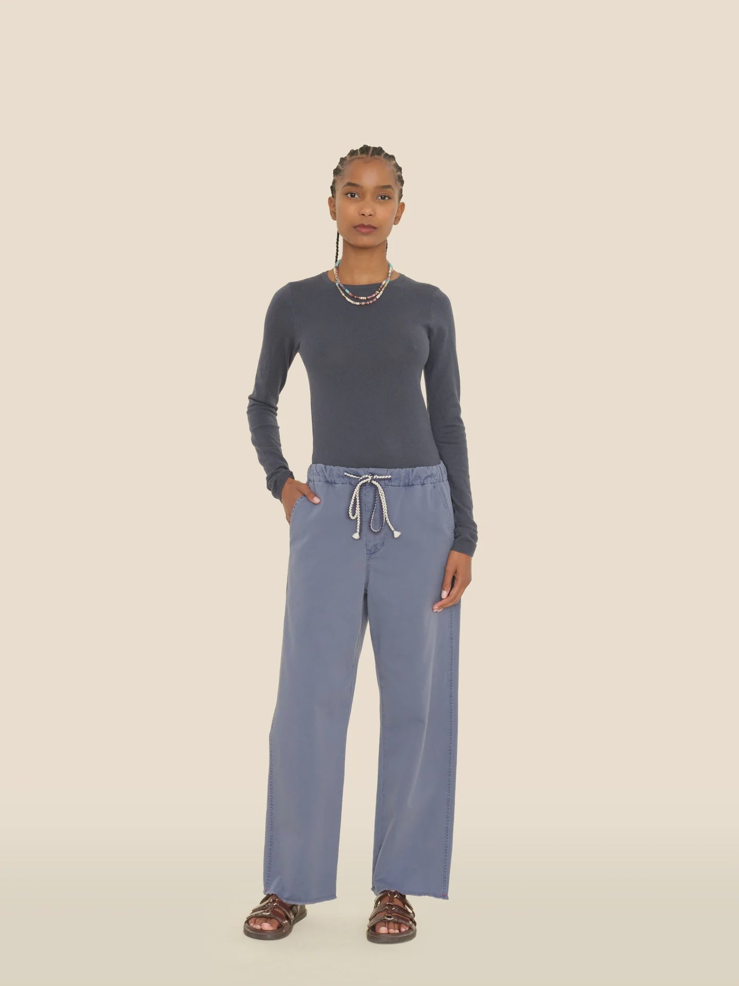 A person stands against a plain background, sporting a dark long-sleeve shirt and the Eli Pant by Xirena, which are light blue, vintage washed cotton twill pants featuring an elasticized drawstring waist. They pair the outfit with brown sandals and accessorize with layered necklaces while wearing their hair in short braids.