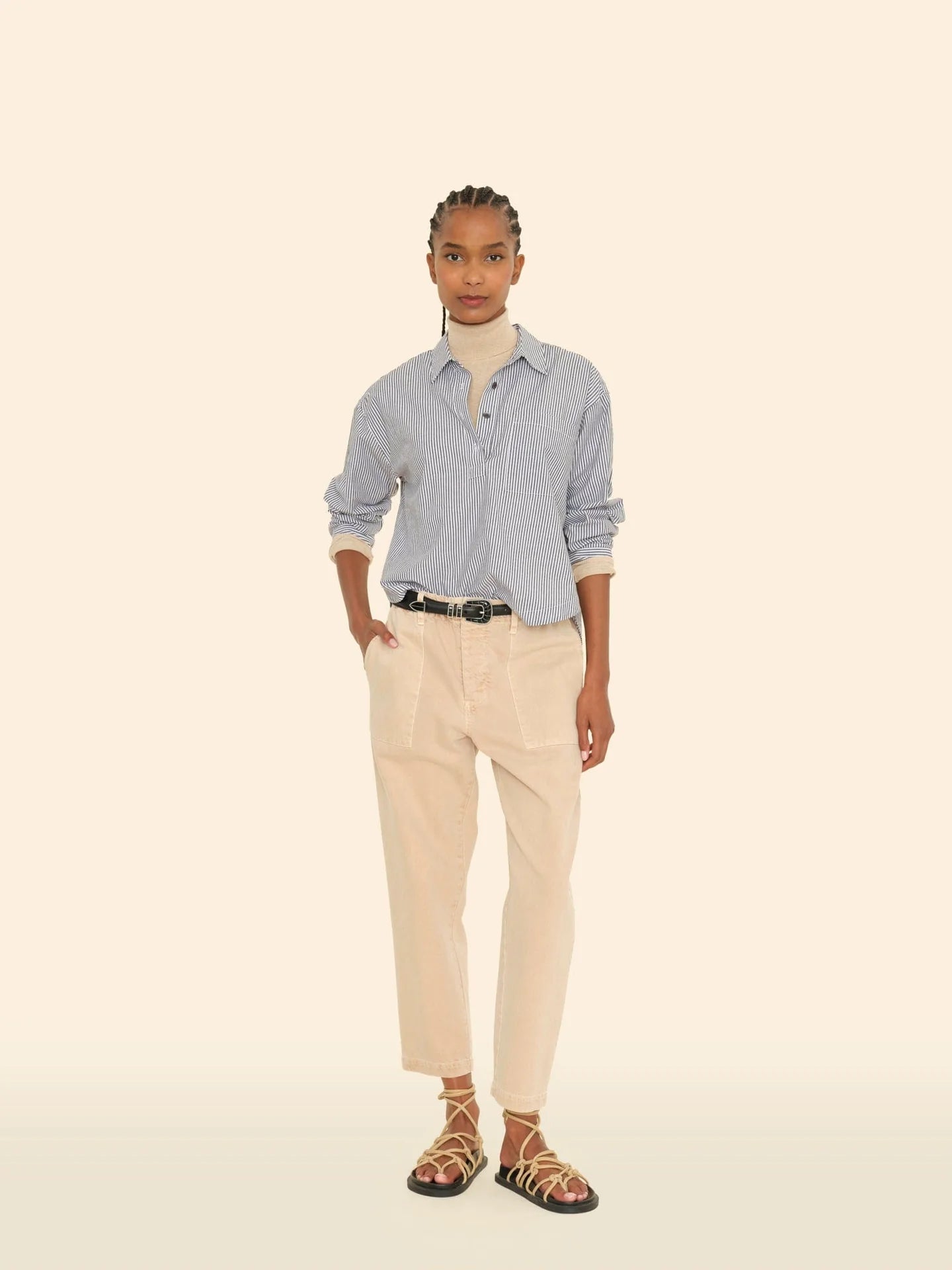 A person stands against a light background wearing a blue button-up shirt and the Mercer Pant by Xirena, featuring beige fabric with patch pockets and a button fly. Black sandals with tan straps complement their braided hair and black belt, while their hands casually rest in the pockets of these recycled denim pants.