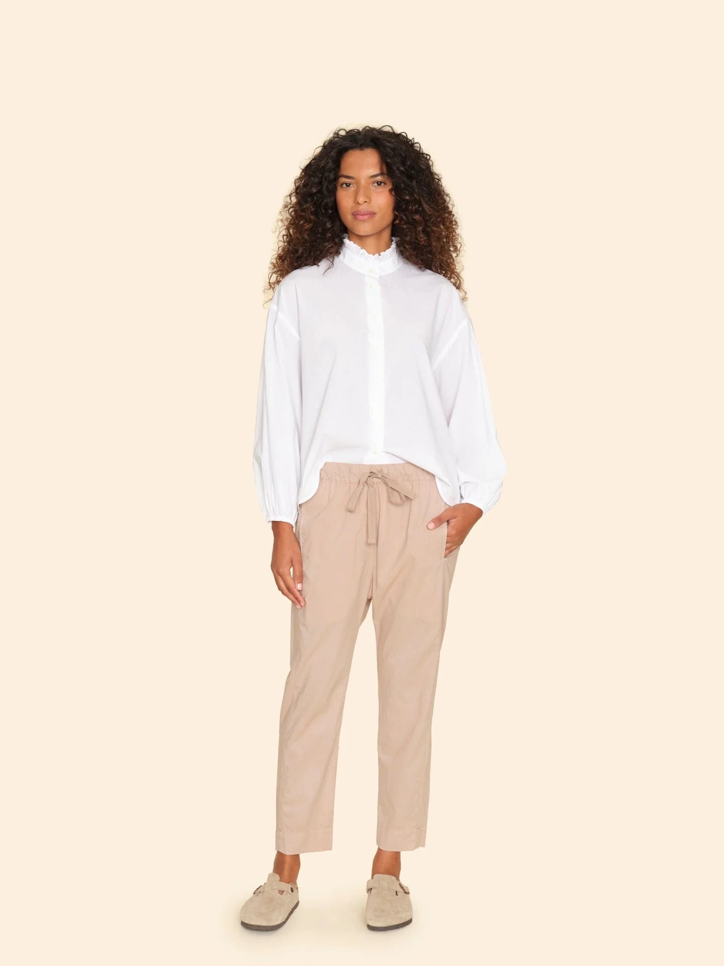 A person with curly hair is wearing the Draper Pant by Xirena, featuring subtle side seam pockets. They style the beige pants with a white blouse and light-colored slip-on shoes, and are standing against a plain beige background, casually resting one hand in their pocket.