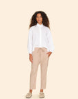 A person with curly hair is wearing the Draper Pant by Xirena, featuring subtle side seam pockets. They style the beige pants with a white blouse and light-colored slip-on shoes, and are standing against a plain beige background, casually resting one hand in their pocket.