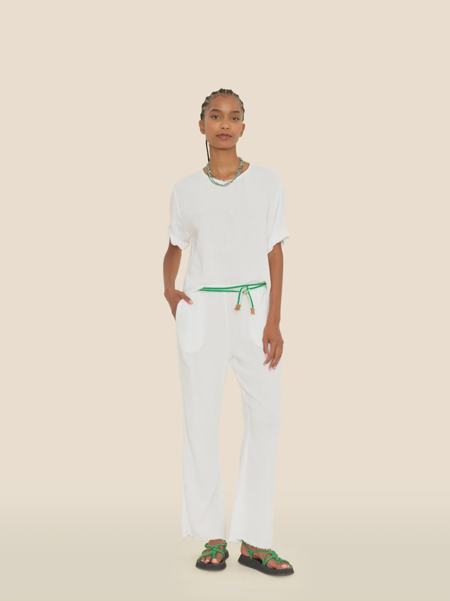 A person dressed in the Bella Pant by Xirena, showcasing an elasticized waist and complemented by a green belt, stands against a light background. Their wide-leg pants effortlessly pair with stylish green sandals. With braided hair, they accessorize seamlessly with a necklace and bracelets, exuding a chic yet relaxed vibe.