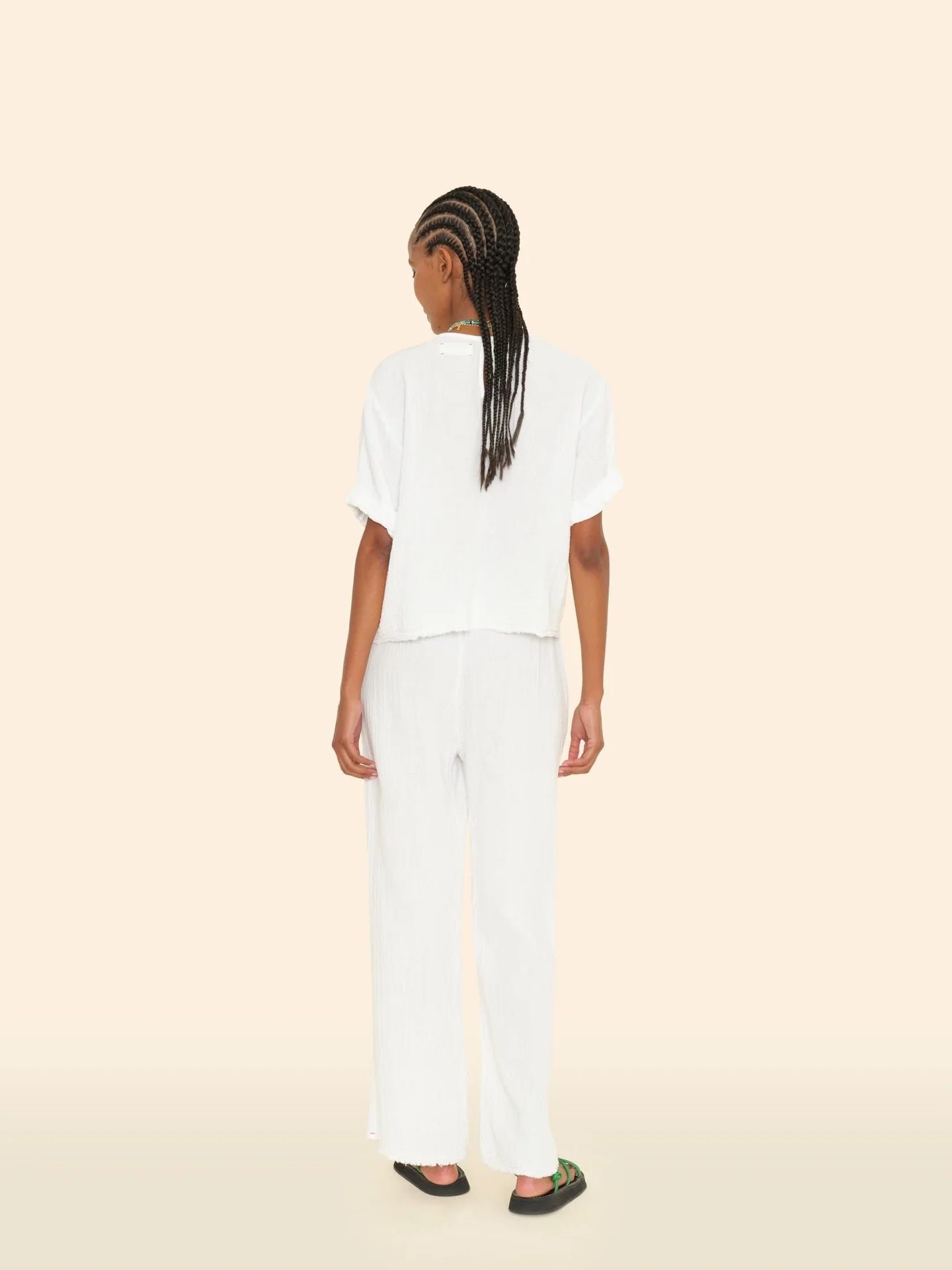 A person with long braided hair stands facing away, wearing a comfortable white short-sleeved top and the Bella Pant by Xirena, designed with loose wide legs and an elasticized waist. They are in black sandals, set against a plain beige background.