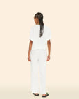 A person with long braided hair stands facing away, wearing a comfortable white short-sleeved top and the Bella Pant by Xirena, designed with loose wide legs and an elasticized waist. They are in black sandals, set against a plain beige background.