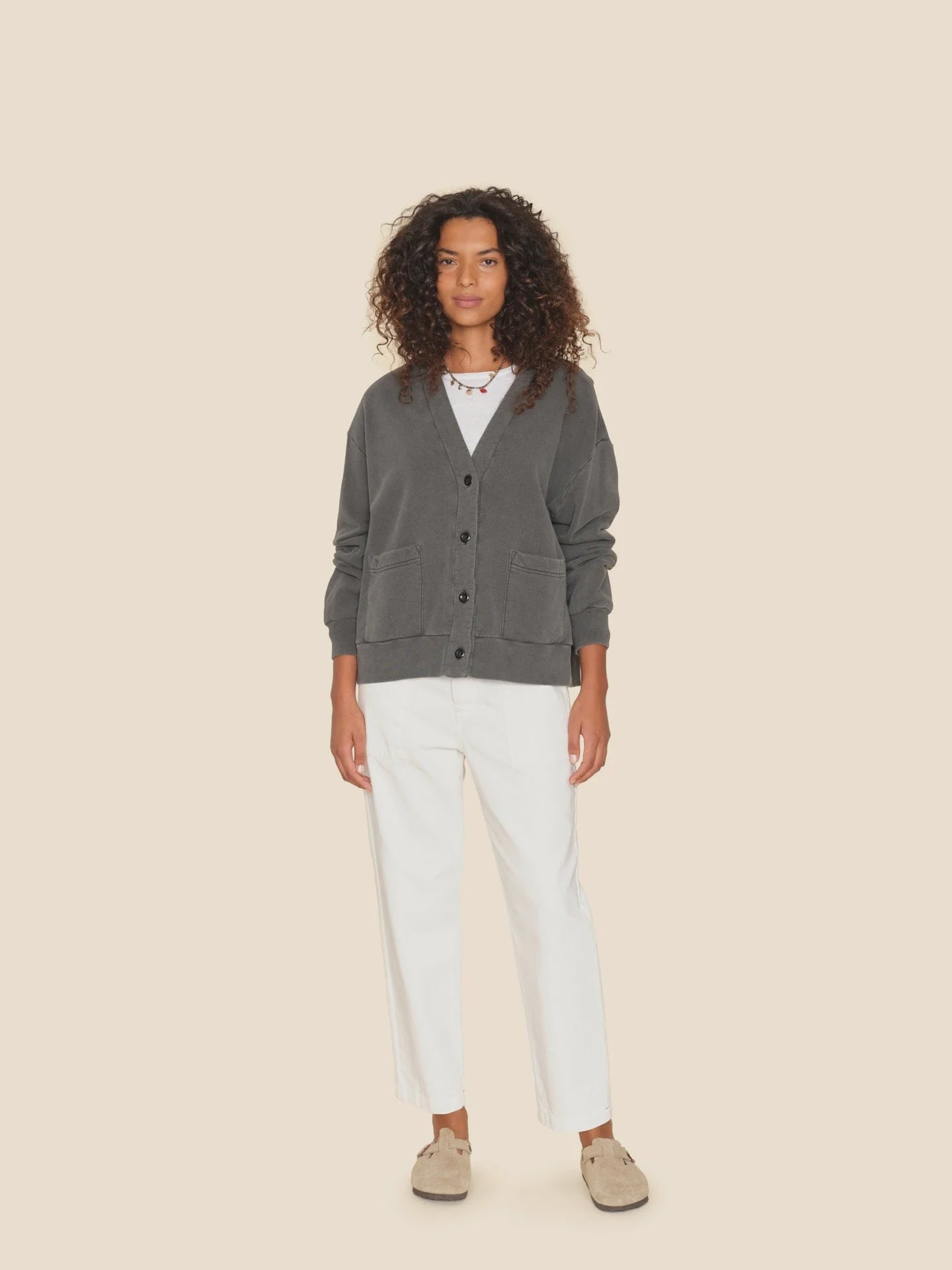 A person with curly hair stands against a beige background, wearing a gray buttoned cardigan over a white shirt and the Mercer Pant from Xirena, crafted from midweight recycled denim. Beige shoes complete the outfit, as the individual looks directly at the camera with a neutral expression.