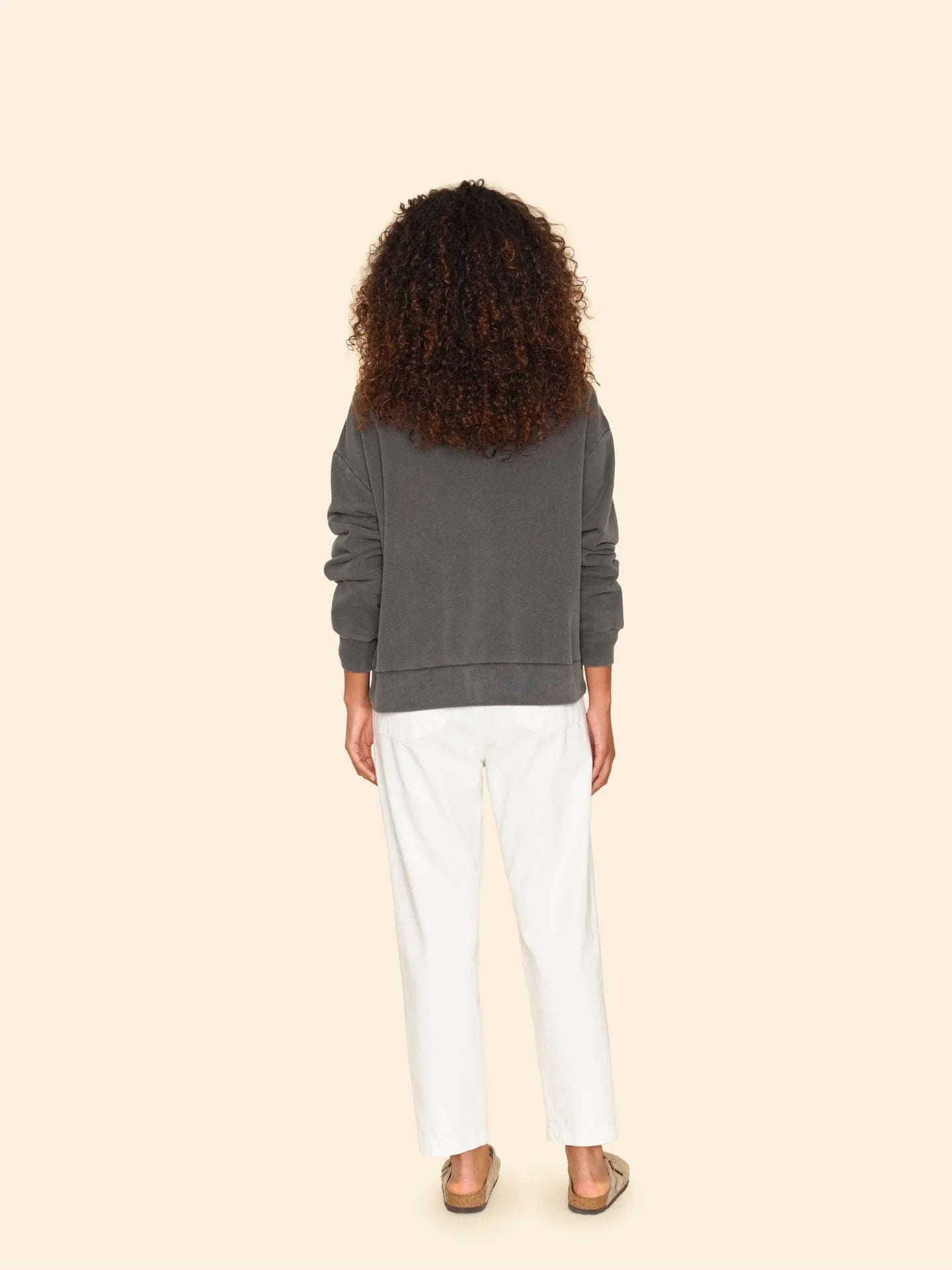 A person with curly hair stands facing away, wearing a dark gray sweatshirt and the Mercer Pant by Xirena, made from white recycled denim with a button fly. They complete the look with sandals against a plain beige background.