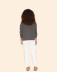 A person with curly hair stands facing away, wearing a dark gray sweatshirt and the Mercer Pant by Xirena, made from white recycled denim with a button fly. They complete the look with sandals against a plain beige background.