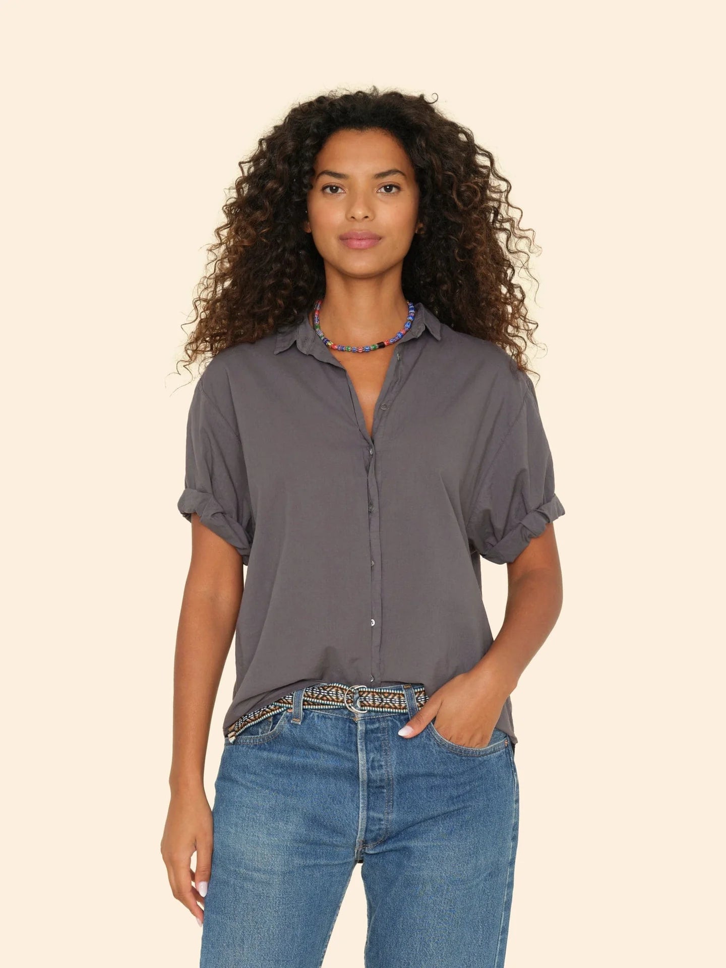 A person with curly hair is wearing a dark, short-sleeved Channing Shirt by Xirena, made from lightweight woven cotton poplin. The shirt is neatly tucked into blue jeans and complemented by a colorful necklace, all set against a plain beige background.