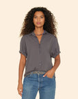 A person with curly hair is wearing a dark, short-sleeved Channing Shirt by Xirena, made from lightweight woven cotton poplin. The shirt is neatly tucked into blue jeans and complemented by a colorful necklace, all set against a plain beige background.