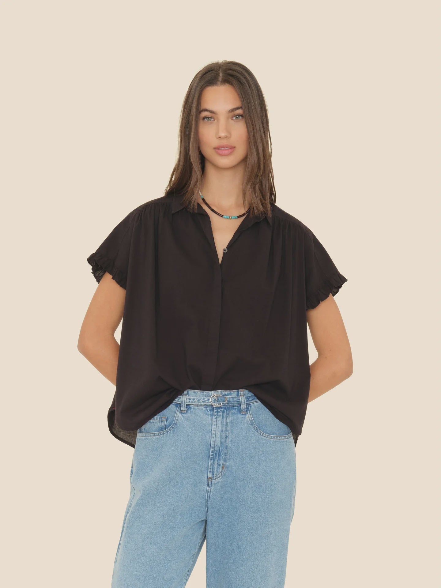 A person with long hair stands against a plain background, wearing the black Noah Shirt by Xirena, featuring ruffle sleeves, paired with light blue jeans. They have their hands in their pockets, a calm expression on their face, and are accessorized with a layered necklace.