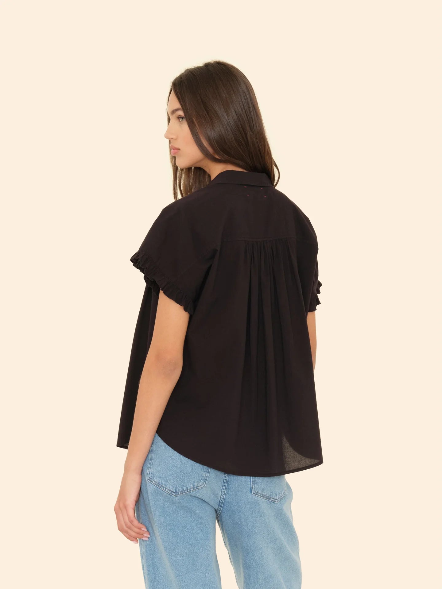 A person with long brown hair is wearing the Noah Shirt by Xirena, a button-front blouse featuring ruffle sleeves, paired with light blue jeans. They are standing against a plain beige background, facing slightly away from the camera.