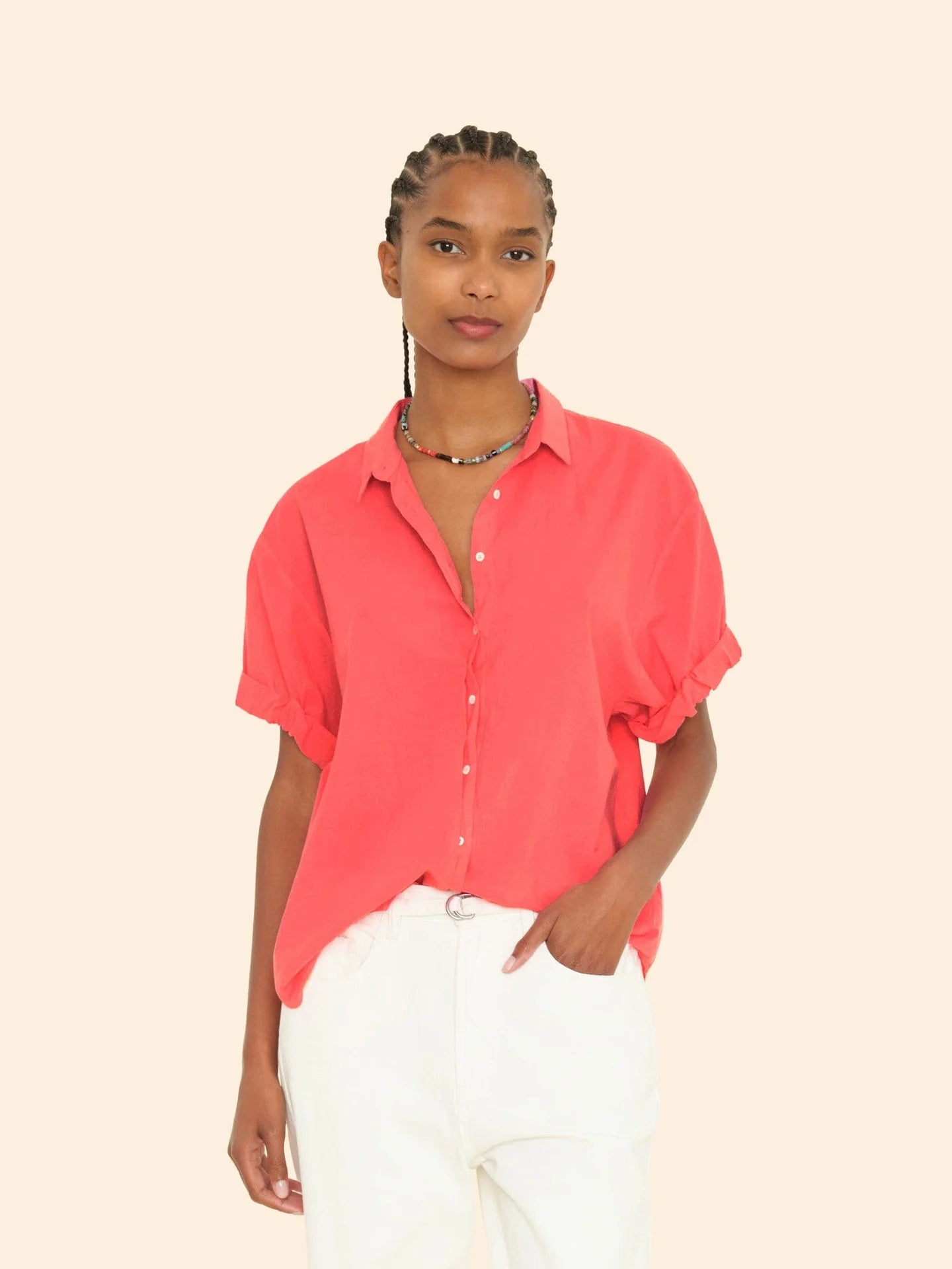 A person in a bright red Channing Shirt by Xirena with rolled sleeves and white pants stands against a cream background. Their braided hair pairs well with a multicolored beaded necklace, highlighting the shirt's boy meets girl vibe and lightweight woven cotton fabric.