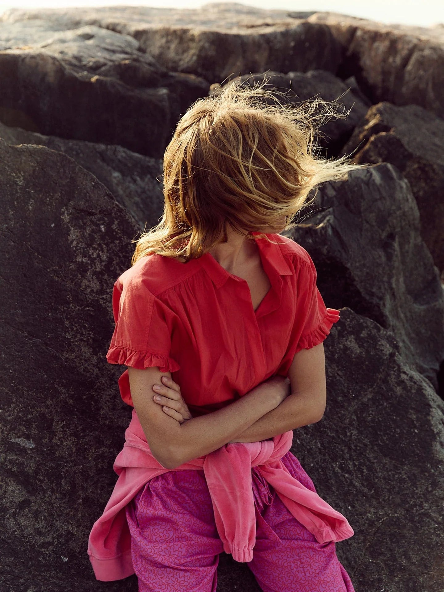 A person with light brown hair, windblown across their face, sits with arms crossed in front of large rocks. They're wearing the red Noah Shirt by Xirena and pink pants.