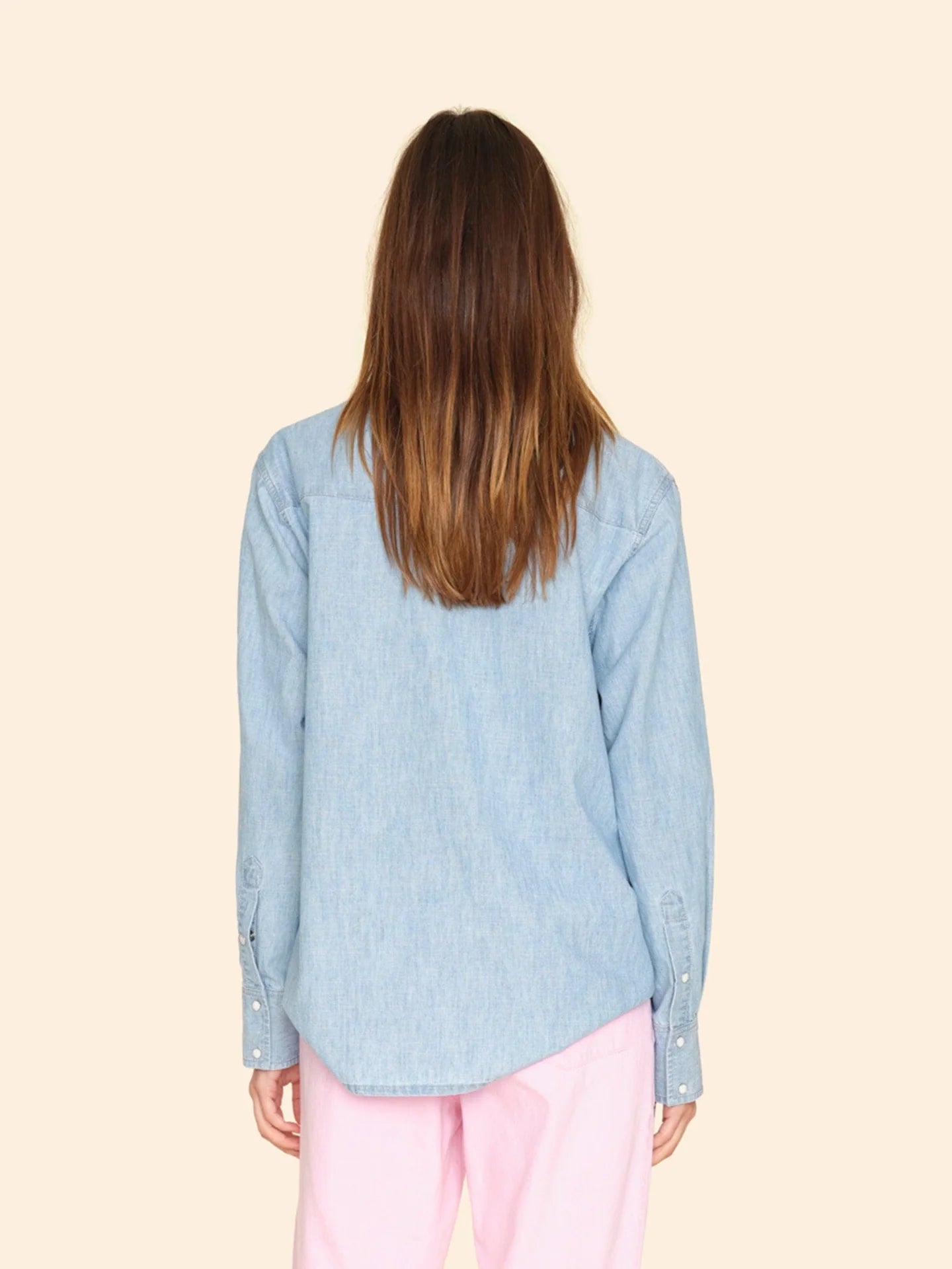 Back view of a person with long brown hair wearing the Griffen Shirt, a Western-inspired light blue denim button-front top by Xirena, paired with pink pants. They stand against a plain beige background.