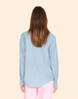 Back view of a person with long brown hair wearing the Griffen Shirt, a Western-inspired light blue denim button-front top by Xirena, paired with pink pants. They stand against a plain beige background.