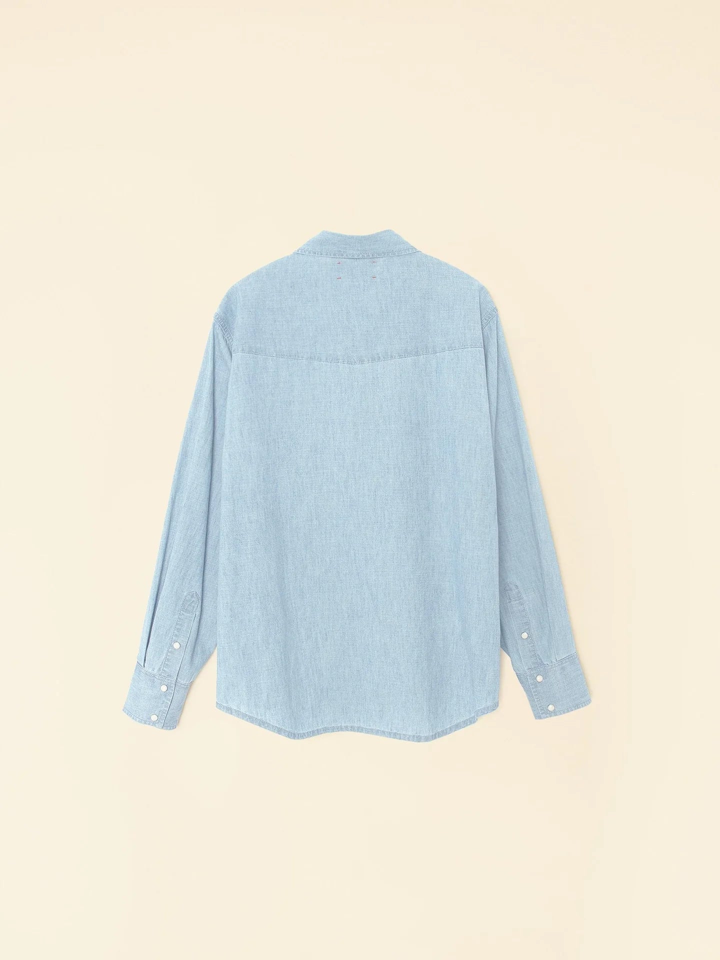 The Griffen Shirt by Xirena, a long-sleeved light blue denim top made from lightweight cotton chambray, is shown from the back against a plain beige background, showcasing its Western-inspired simple design and buttoned cuffs.