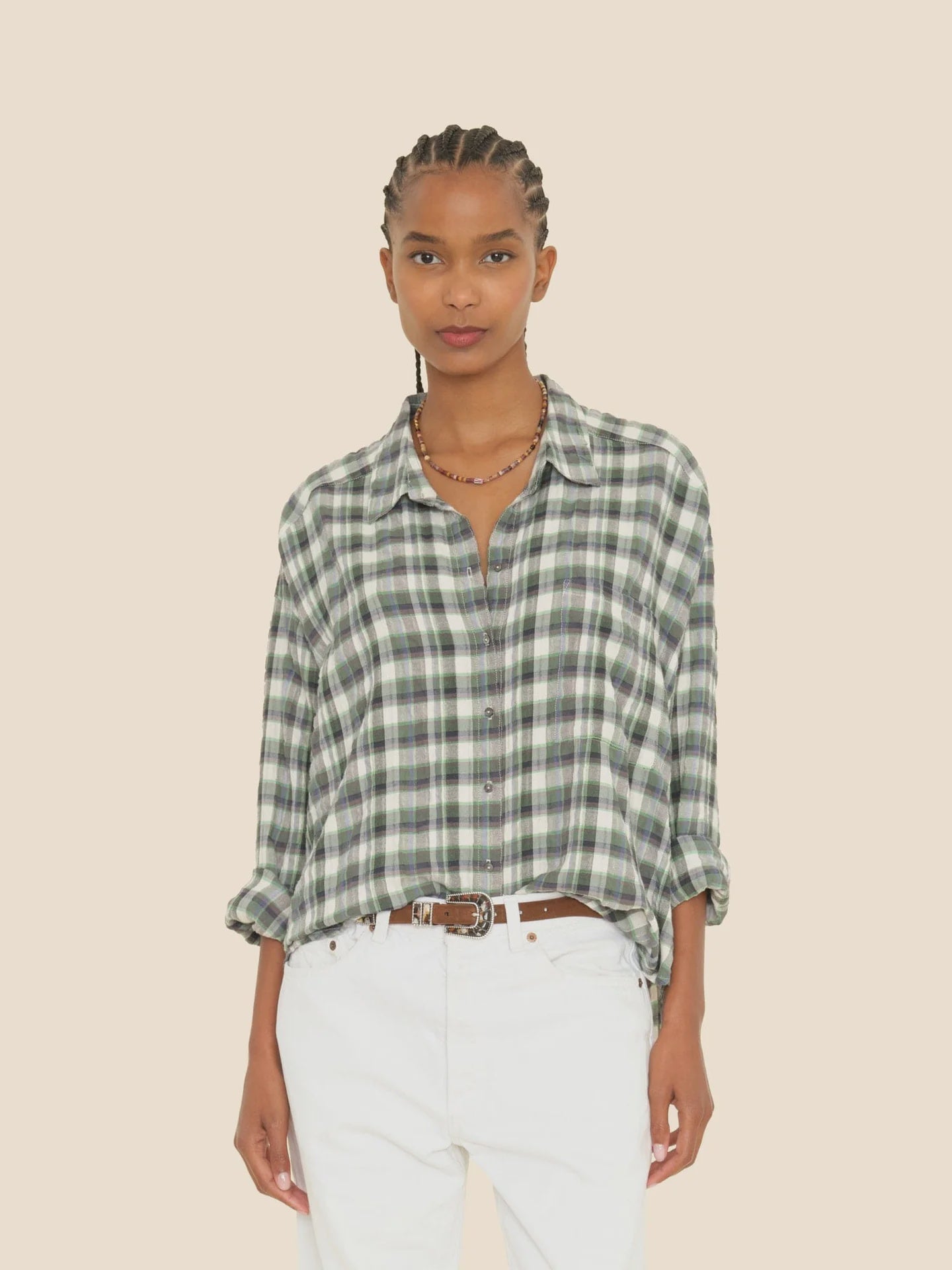 A person with braided hair wears the Jordy Shirt from Xirena, a green and white button-front plaid shirt, paired with white pants against a beige background. The boxy fit of their attire complements their relaxed stance as they have their hands in their pockets.