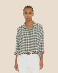 A person with braided hair wears the Jordy Shirt from Xirena, a green and white button-front plaid shirt, paired with white pants against a beige background. The boxy fit of their attire complements their relaxed stance as they have their hands in their pockets.