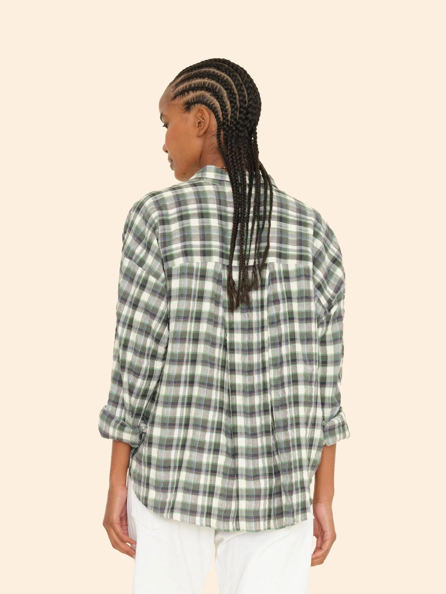 A person with braided hair is wearing the Jordy Shirt, a green and gray plaid shirt with a boxy fit from Xirena, paired with white pants. They stand against a plain beige background, their back facing the camera. The outfit boasts an effortless style made in Los Angeles.