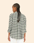 A person with braided hair is wearing the Jordy Shirt, a green and gray plaid shirt with a boxy fit from Xirena, paired with white pants. They stand against a plain beige background, their back facing the camera. The outfit boasts an effortless style made in Los Angeles.