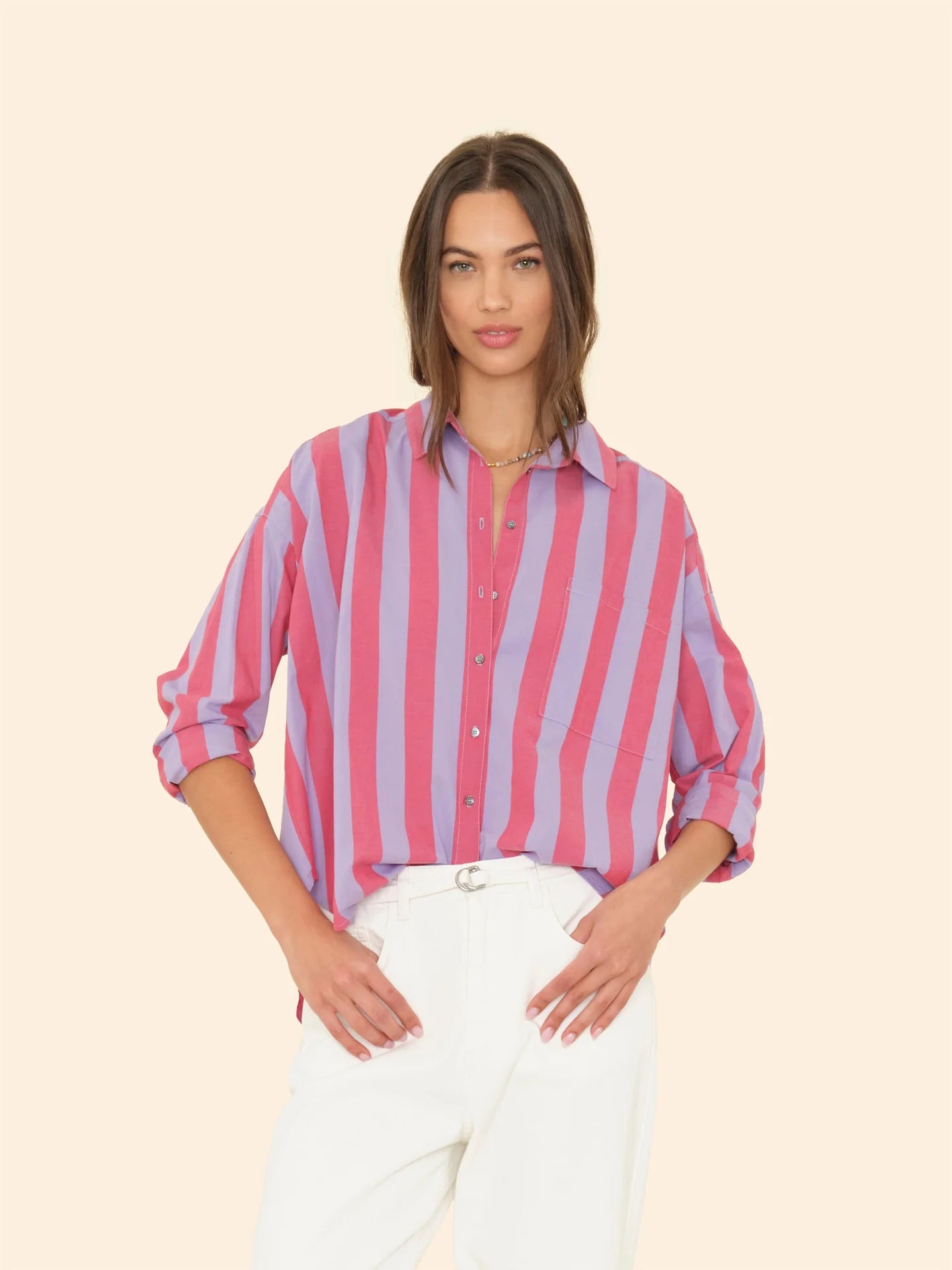 A woman stands wearing the Jordy Shirt by Xirena, featuring a boxy fit and pink and purple stripes, with its button-front design and sleeves casually rolled up. She pairs it with white pants as her long, straight hair cascades while she poses against a plain beige background.