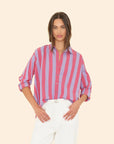 A woman stands wearing the Jordy Shirt by Xirena, featuring a boxy fit and pink and purple stripes, with its button-front design and sleeves casually rolled up. She pairs it with white pants as her long, straight hair cascades while she poses against a plain beige background.