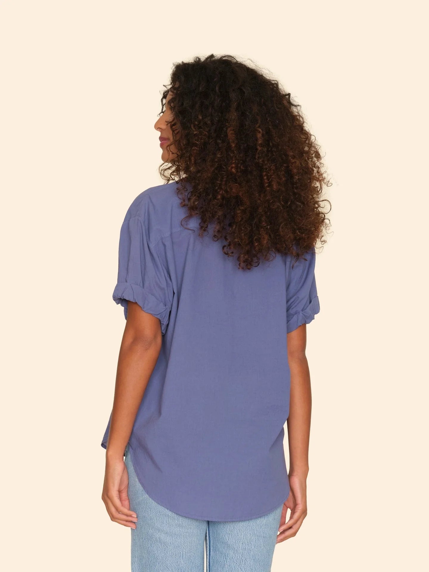A woman with curly hair stands facing away, wearing the Xirena Channing Shirt S25 in purple and light blue jeans against a plain beige background.