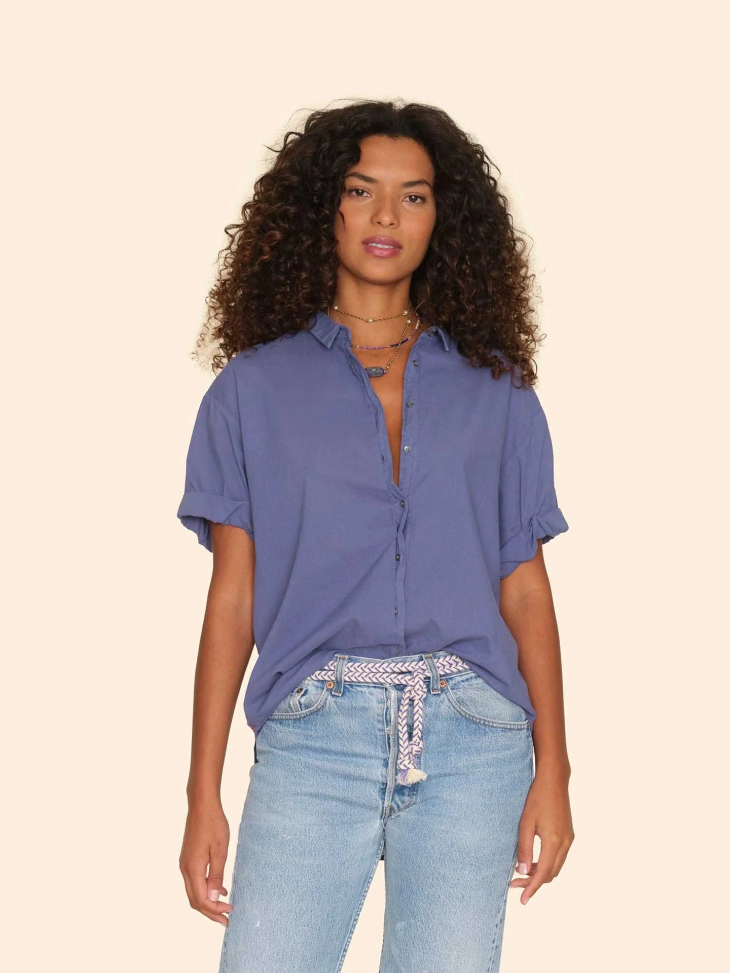 A woman with curly hair wears the Xirena Channing Shirt S25 in lightweight blue chambray, styled with light blue jeans and a woven belt, channeling a &quot;boy meets girl&quot; vibe against a plain beige background.