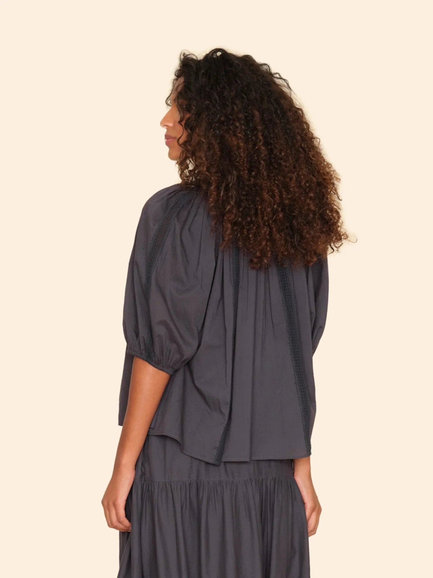 A person with curly hair stands against a plain background, donning the loose-fitting Evynn Shirt by Xirena in a dark color. The shirt features raglan bubble sleeves and lace insets and is crafted from light cotton voile. The person is turned slightly to the side, looking away from the camera.